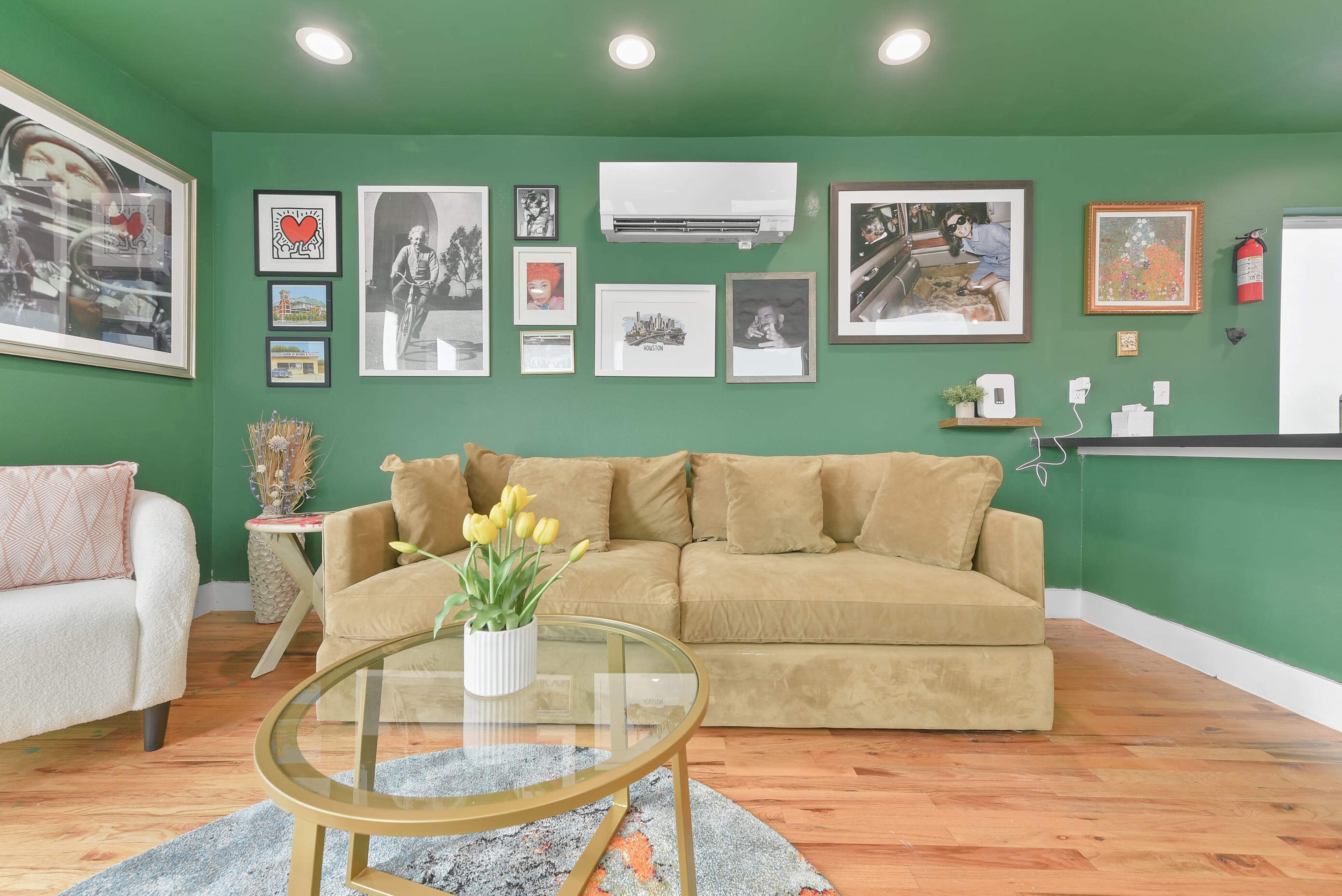 The image shows a cozy living room with a green wall, a beige couch adorned with pillows, a glass coffee table with a vase of yellow tulips, and various framed artworks displayed on the walls.
