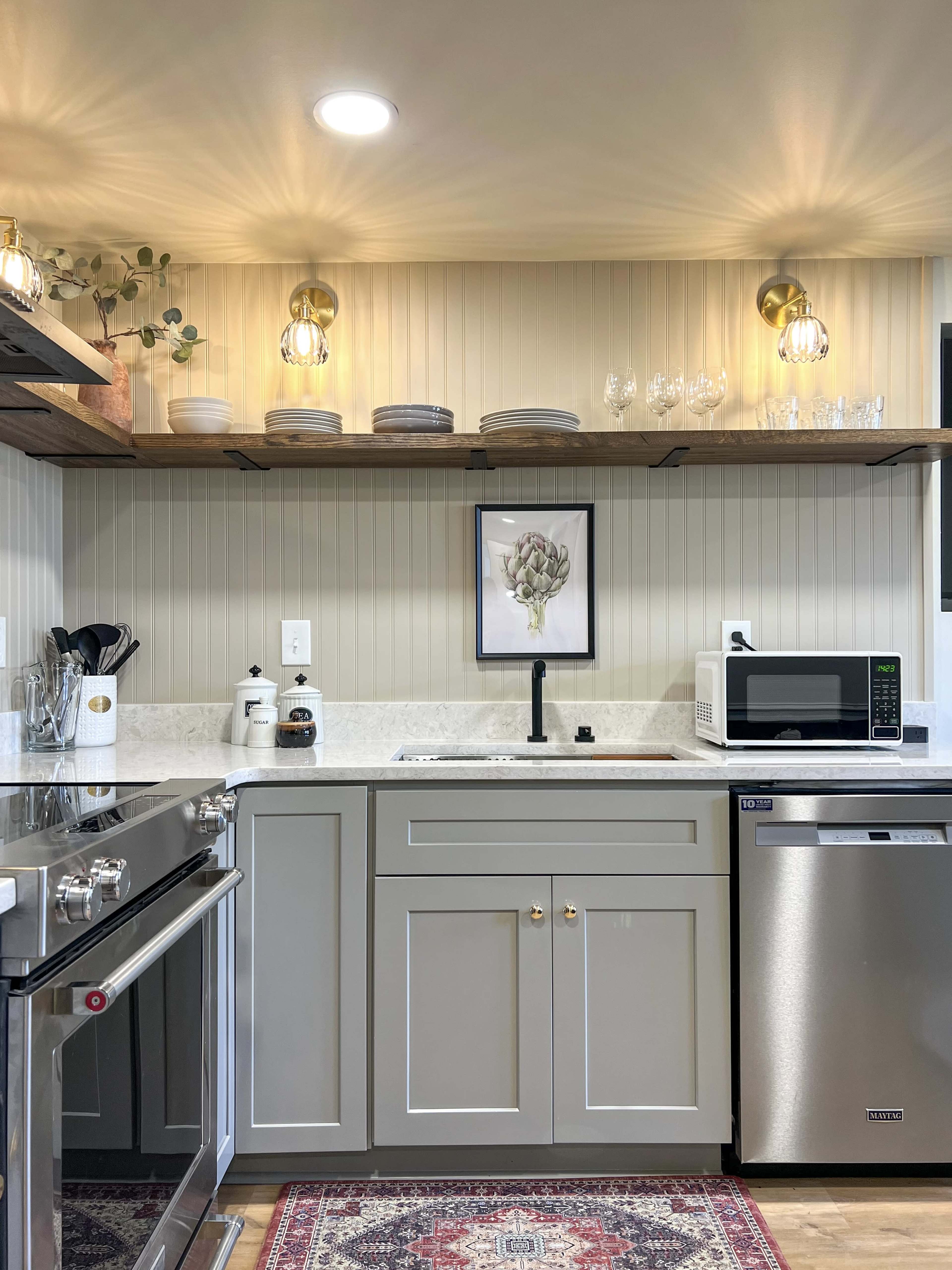 A modern kitchen with a stainless steel stove, a microwave, and open shelving displaying plates and glassware above a white countertop.