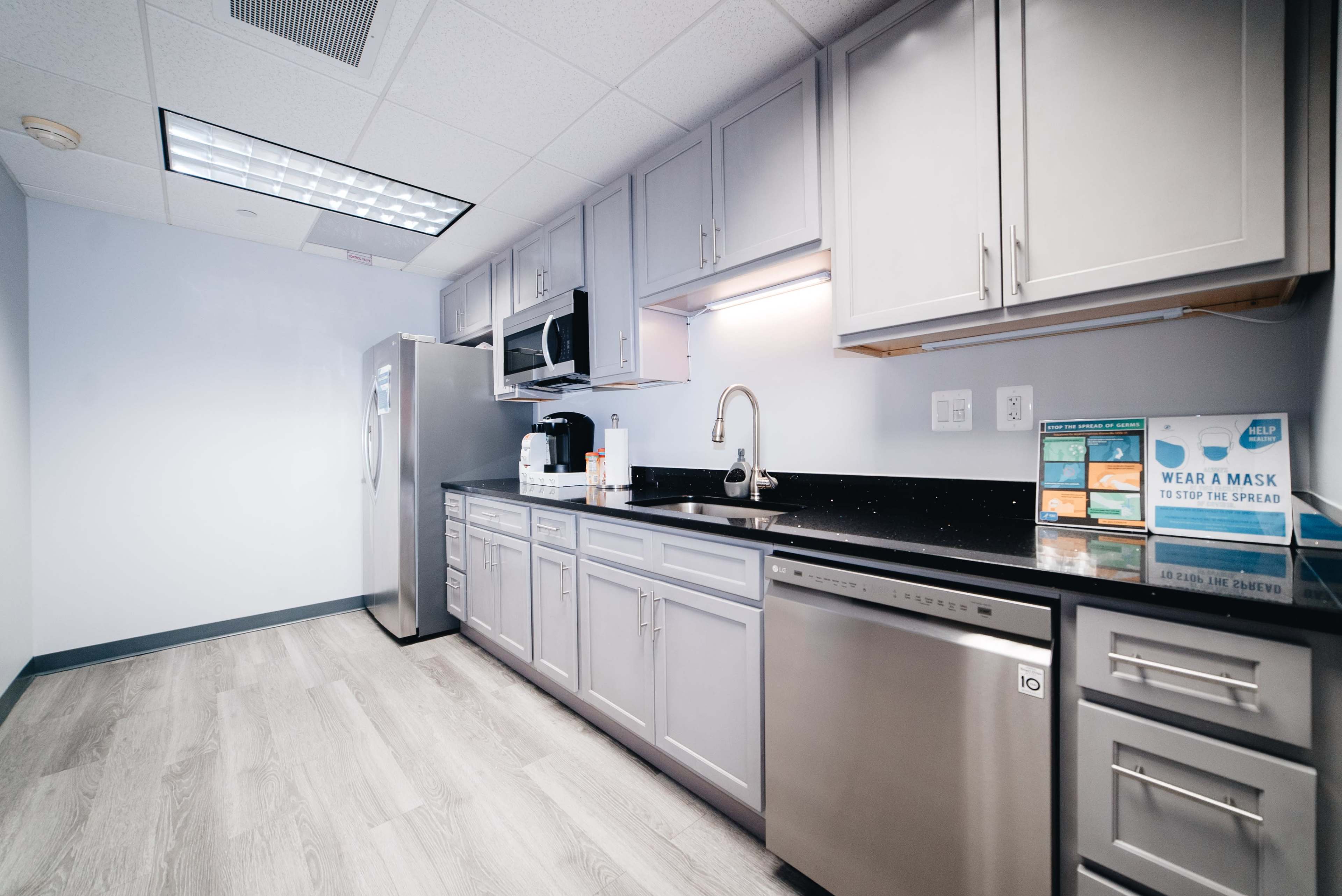 The image shows a modern kitchenette with gray cabinets, a stainless steel refrigerator, a microwave, and a sink, all against a light blue wall.