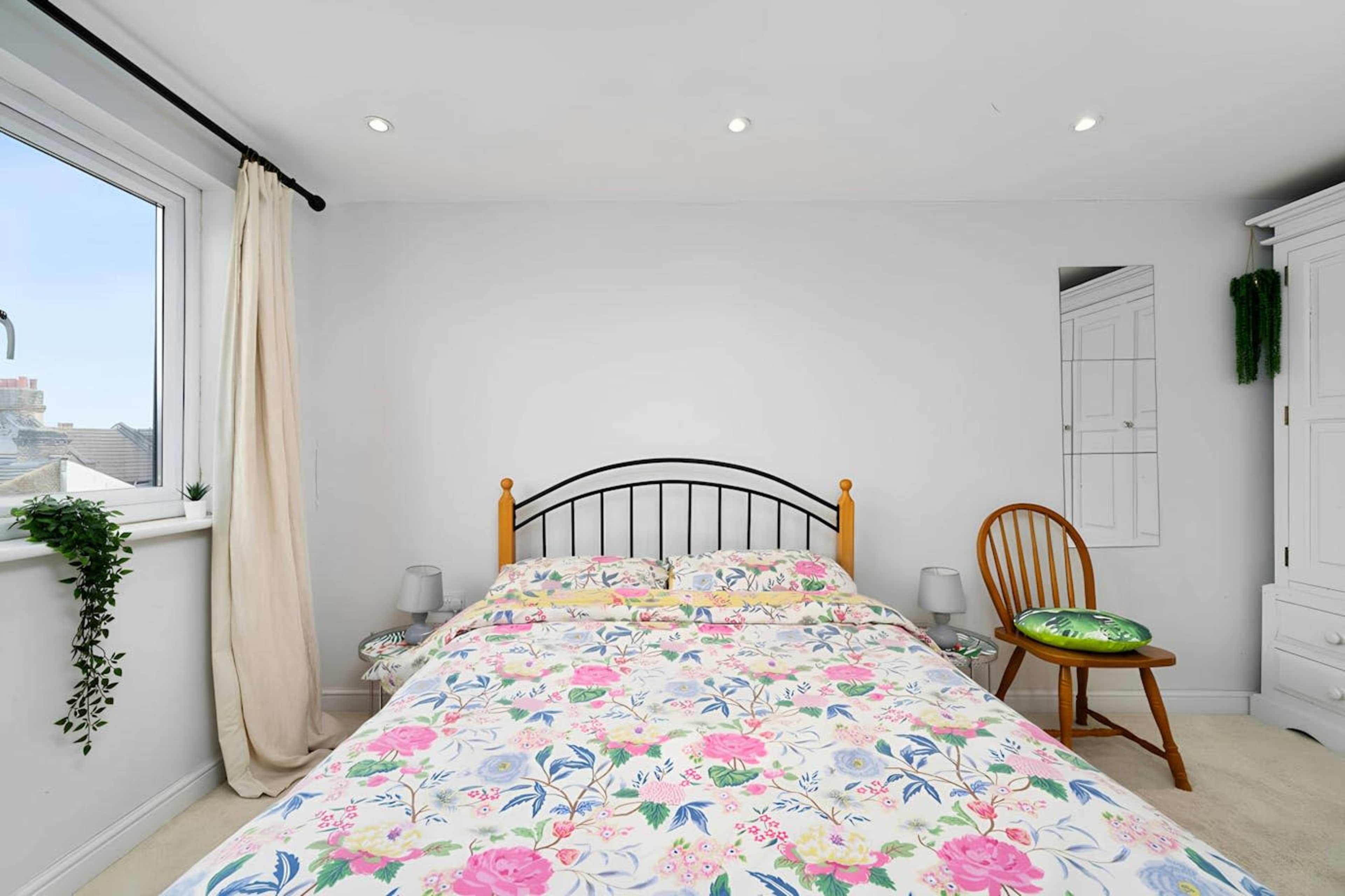 The image shows a bedroom with a double bed adorned in a floral patterned duvet, a wooden chair, a white wardrobe, and a window allowing natural light.