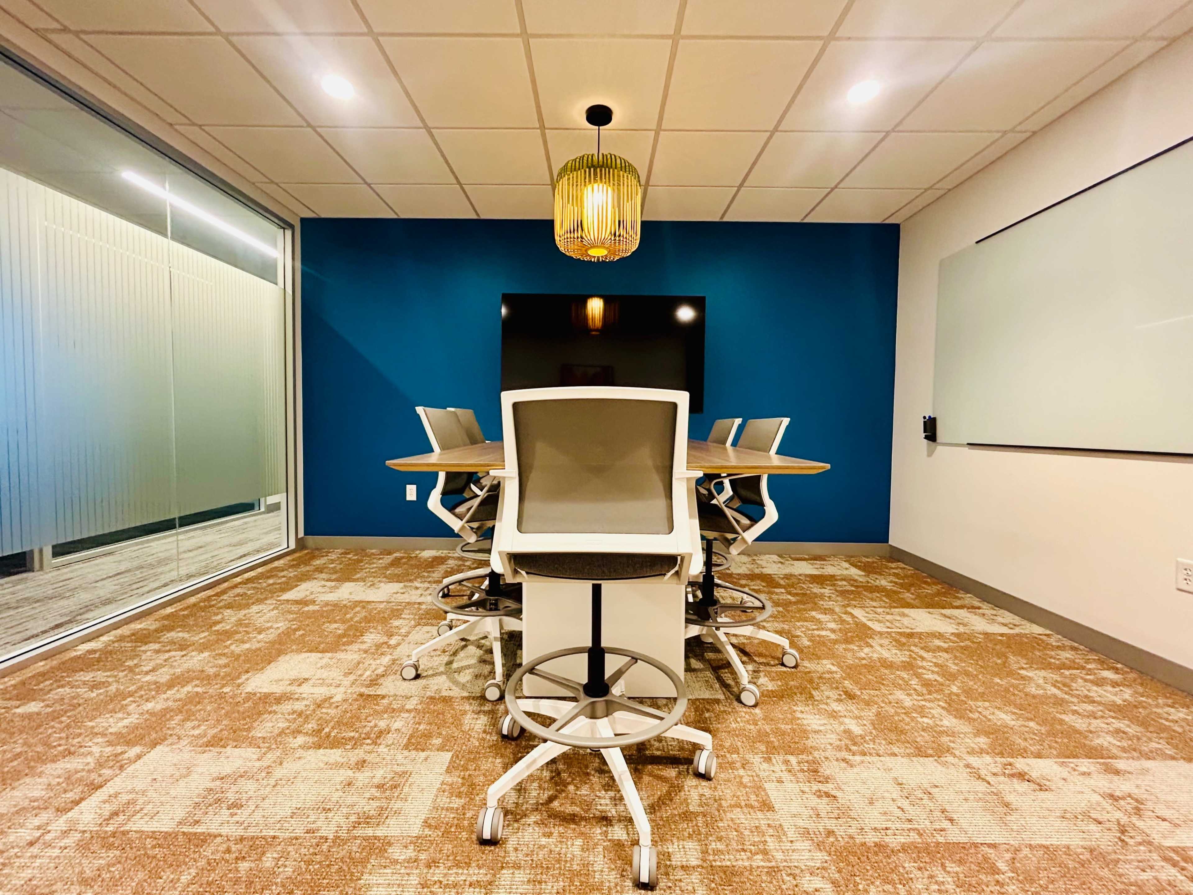 A modern conference room features a long wooden table surrounded by several ergonomic chairs, with a large screen on the wall and a whiteboard.