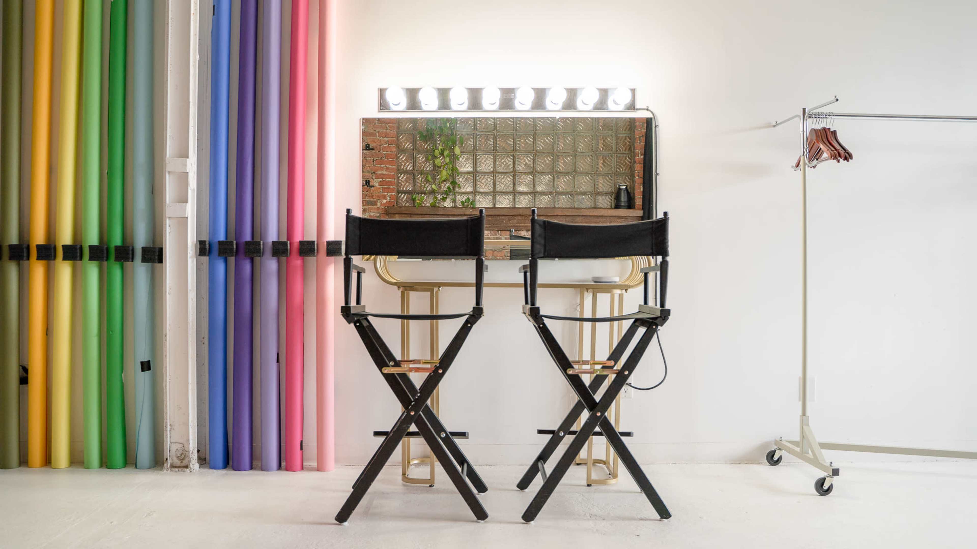 The image shows two black director's chairs in front of a mirrored vanity with lights, flanked by a backdrop of colorful vertical rolls of paper on one side and a clothing rack on the other.