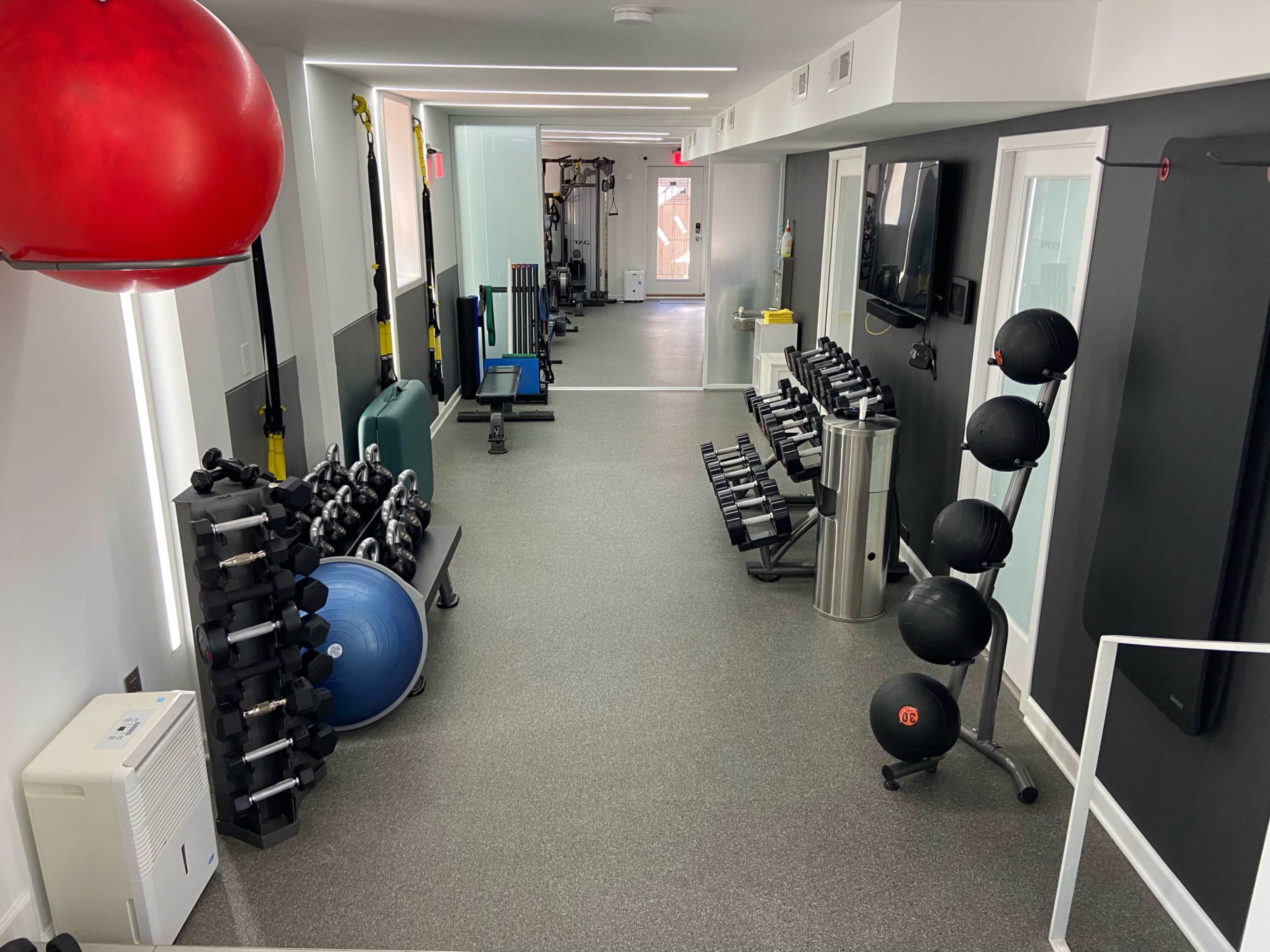 A fitness gym with various equipment, including dumbbells, a stability ball, and a rowing machine, arranged along the walls of a well-lit room.