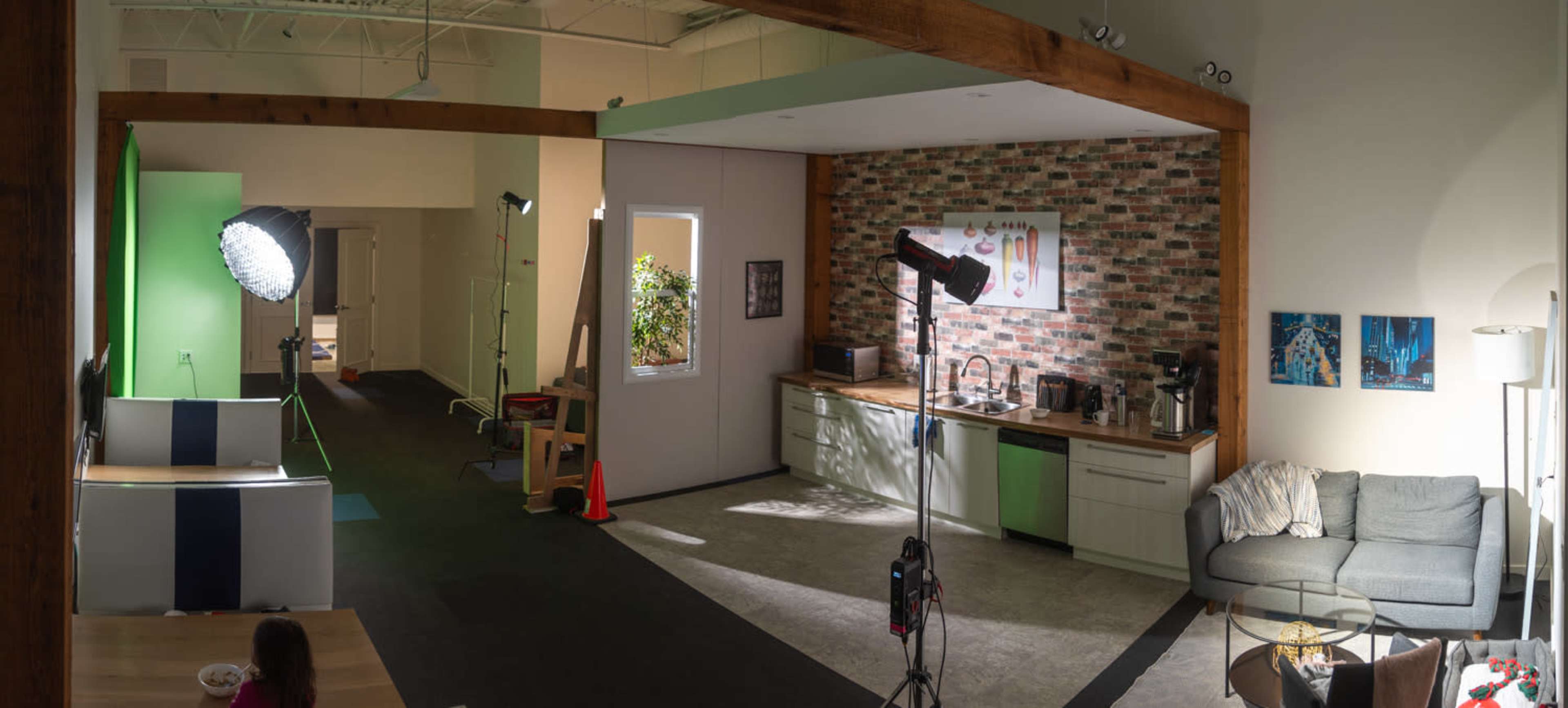 A spacious interior featuring a kitchen area with a brick accent wall, a seating area with a couch, and studio lighting equipment set up for a photo or video shoot.