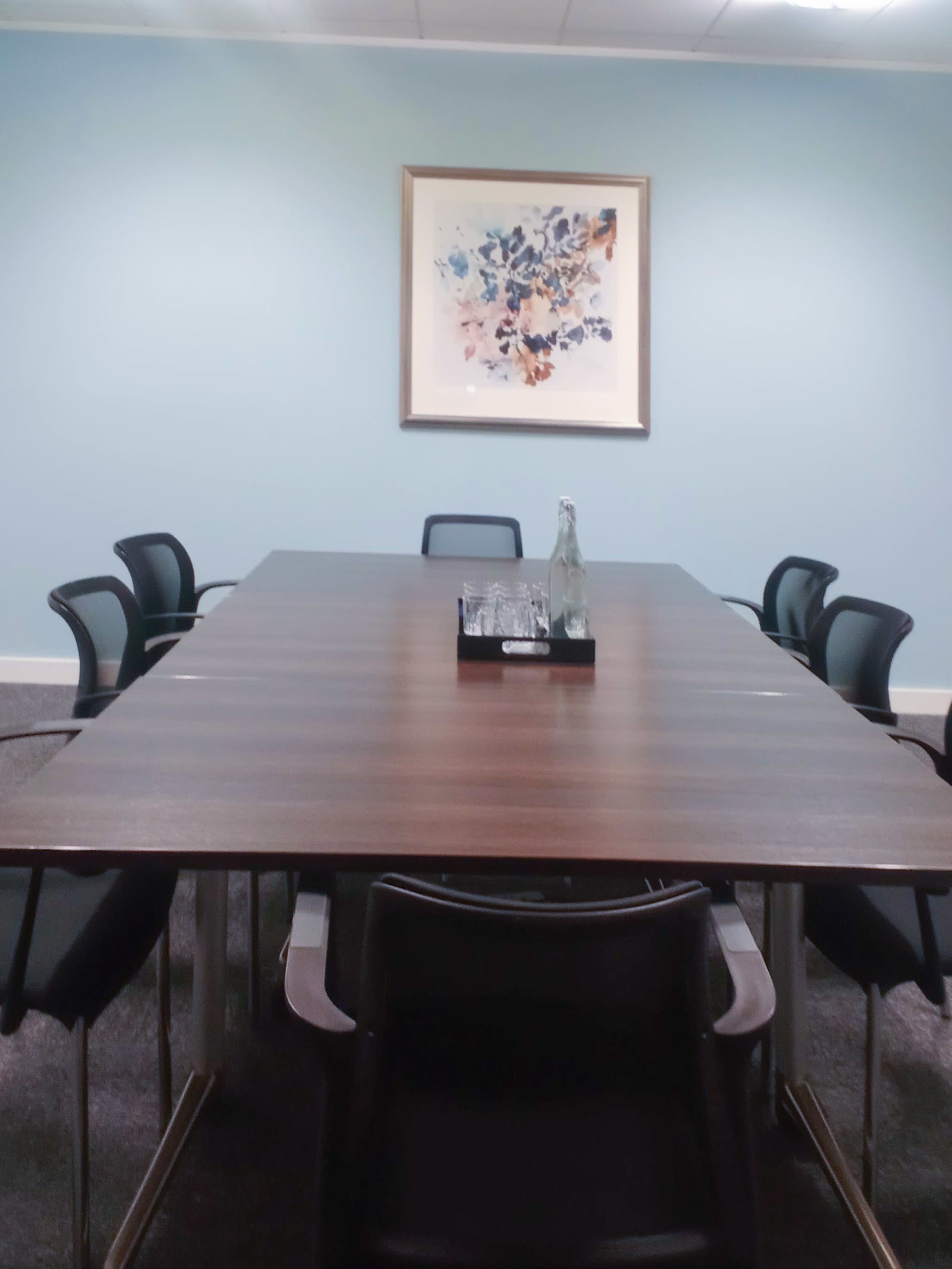 A long, wooden conference table is set with a water bottle and glasses, surrounded by black chairs in a light blue-walled meeting room.