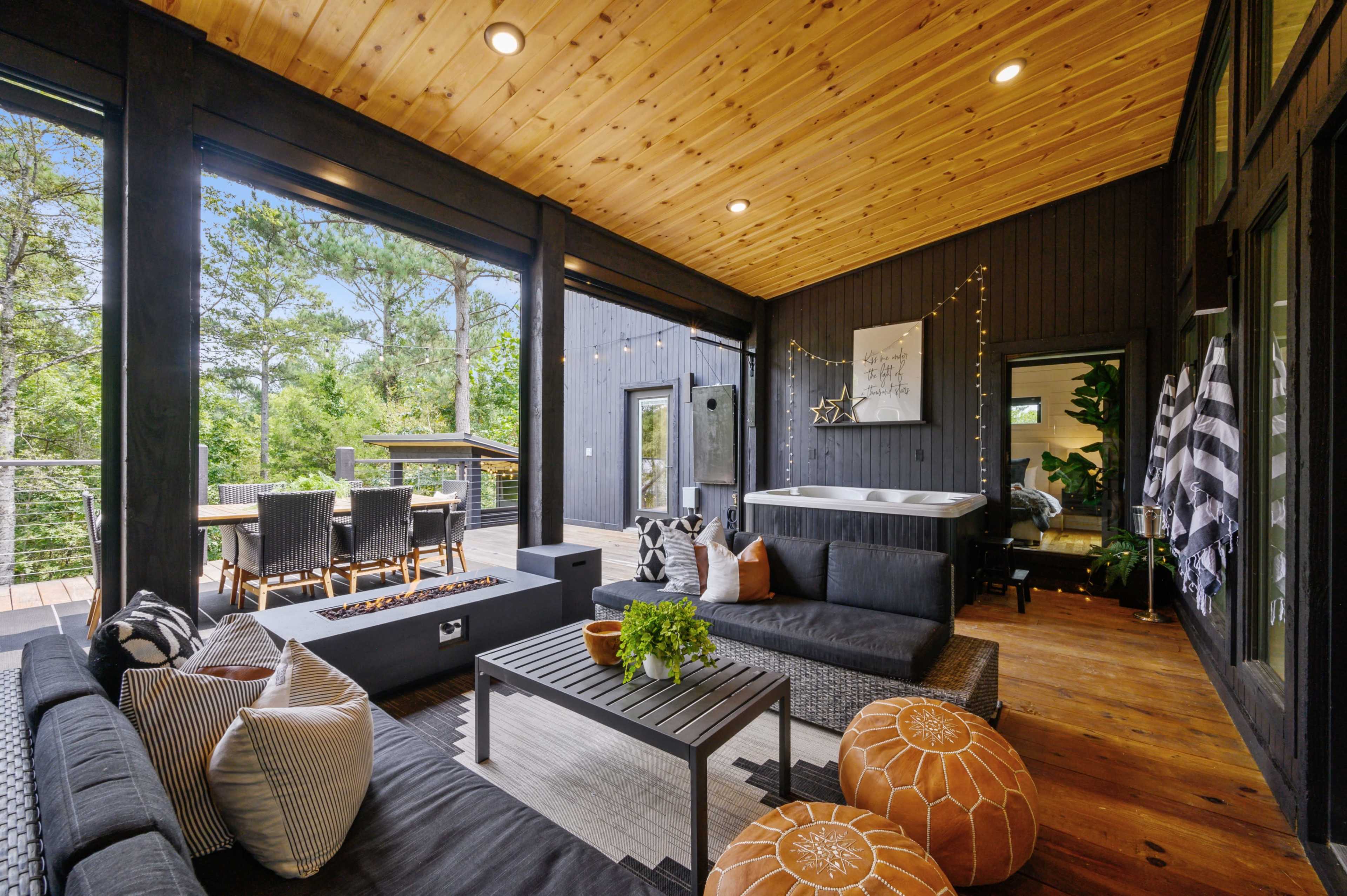 A covered outdoor living space featuring a seating area with dark wicker furniture, a hot tub, and a dining table against a backdrop of greenery.