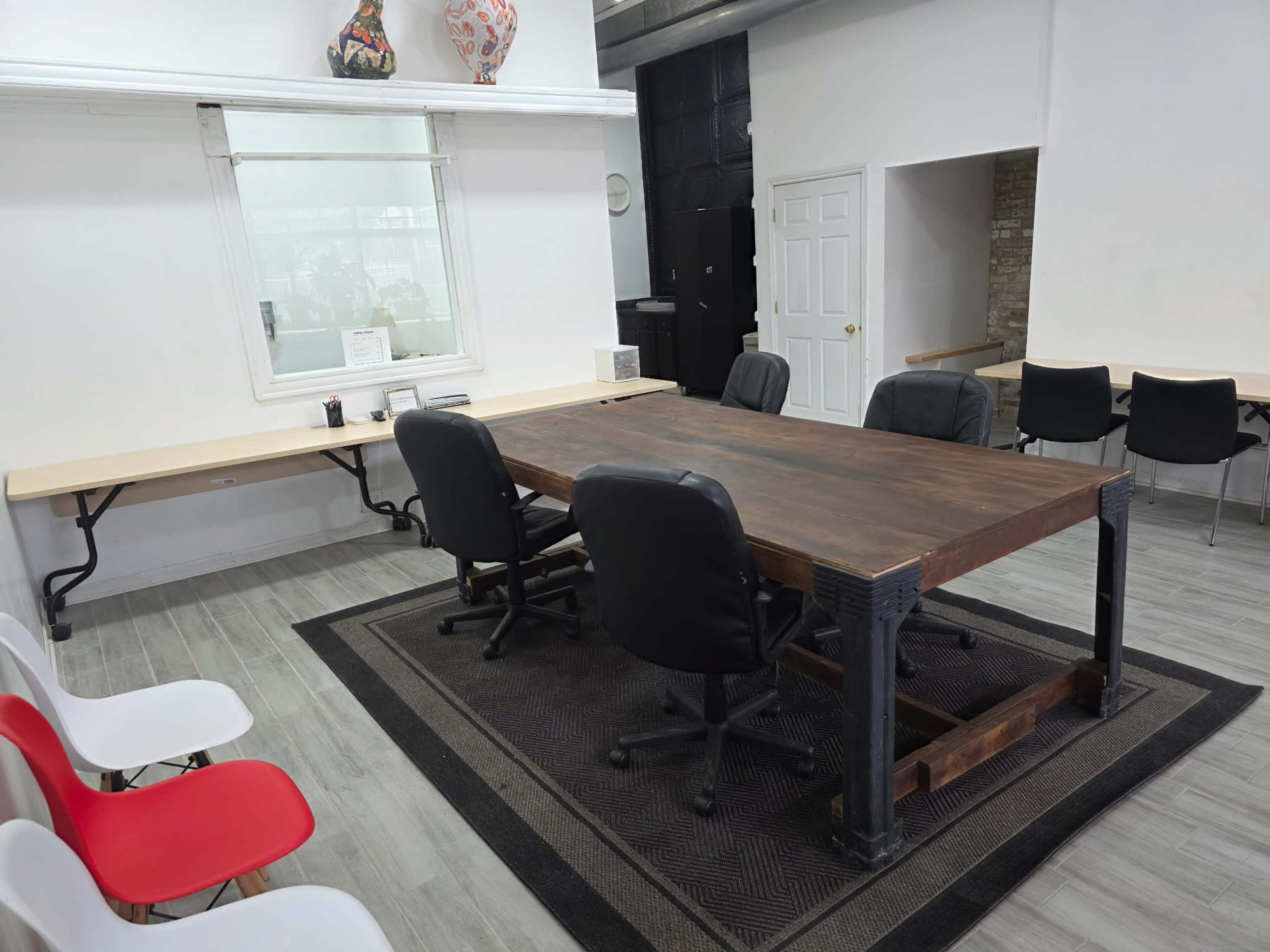 The image shows a meeting room with a large wooden table surrounded by black office chairs, and a few white and red chairs along the side walls.