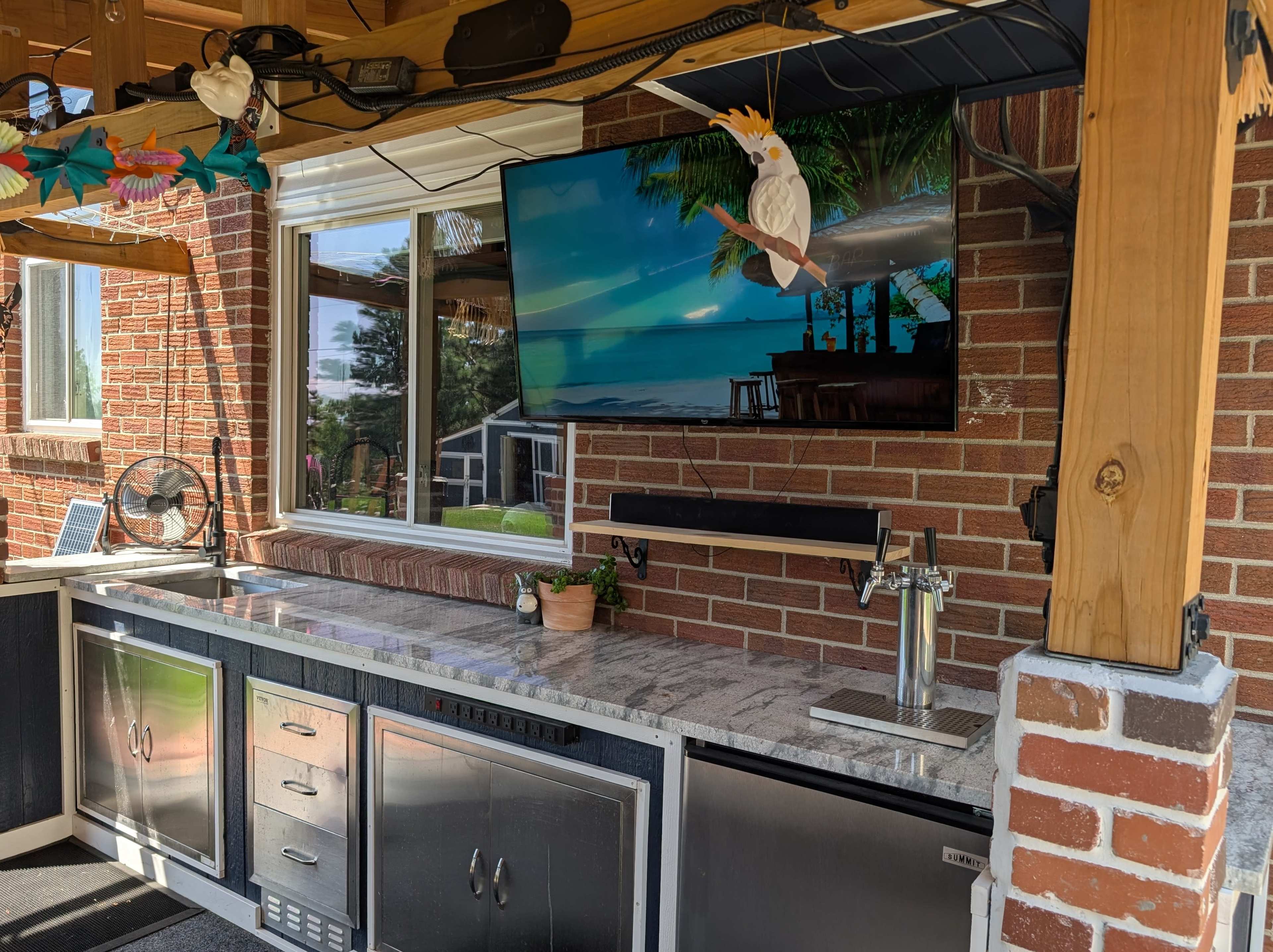 The image shows an outdoor kitchen featuring a granite countertop, stainless steel appliances, and a mounted television displaying a beach scene.