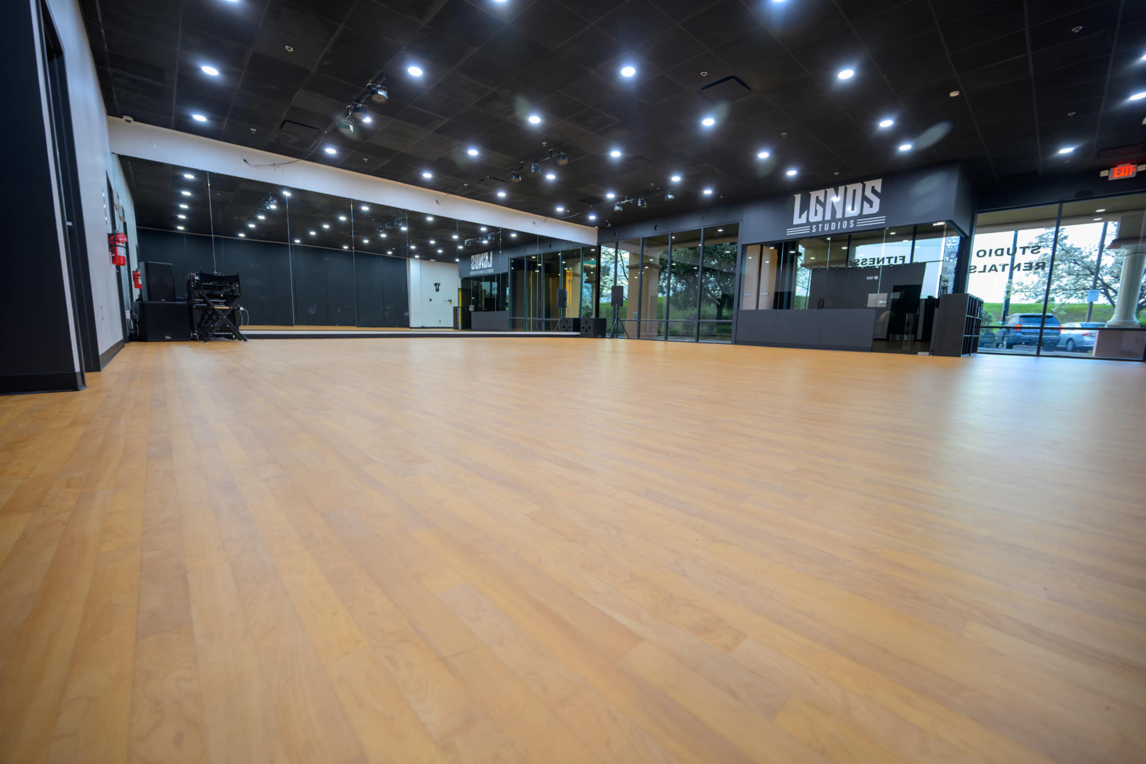 The image shows an empty dance studio with wooden flooring, large mirrors on one wall, and overhead lighting.