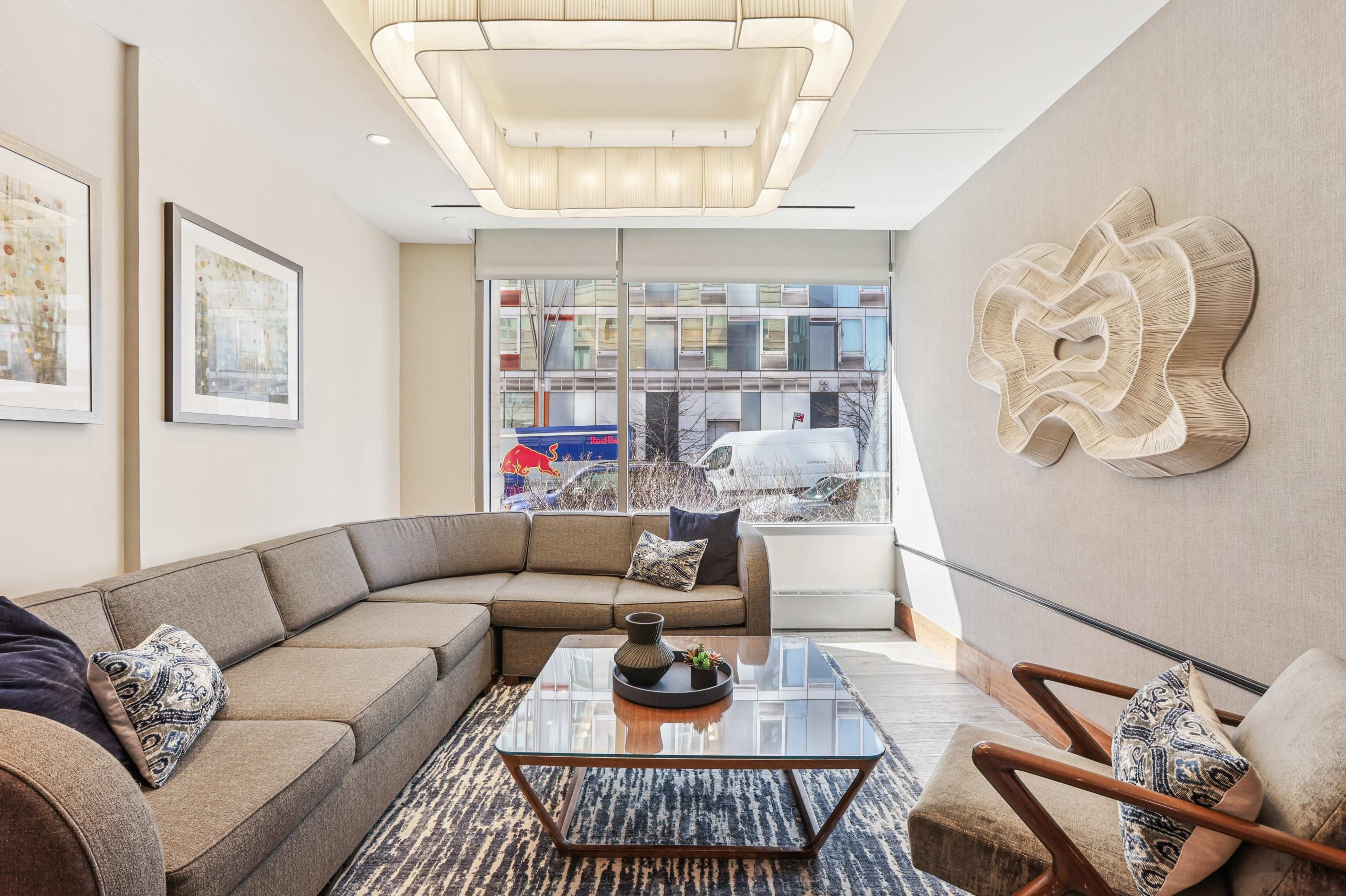 The image shows a modern lounge area with a curved sofa, a glass coffee table, and decorative wall art, illuminated by an overhead light fixture.