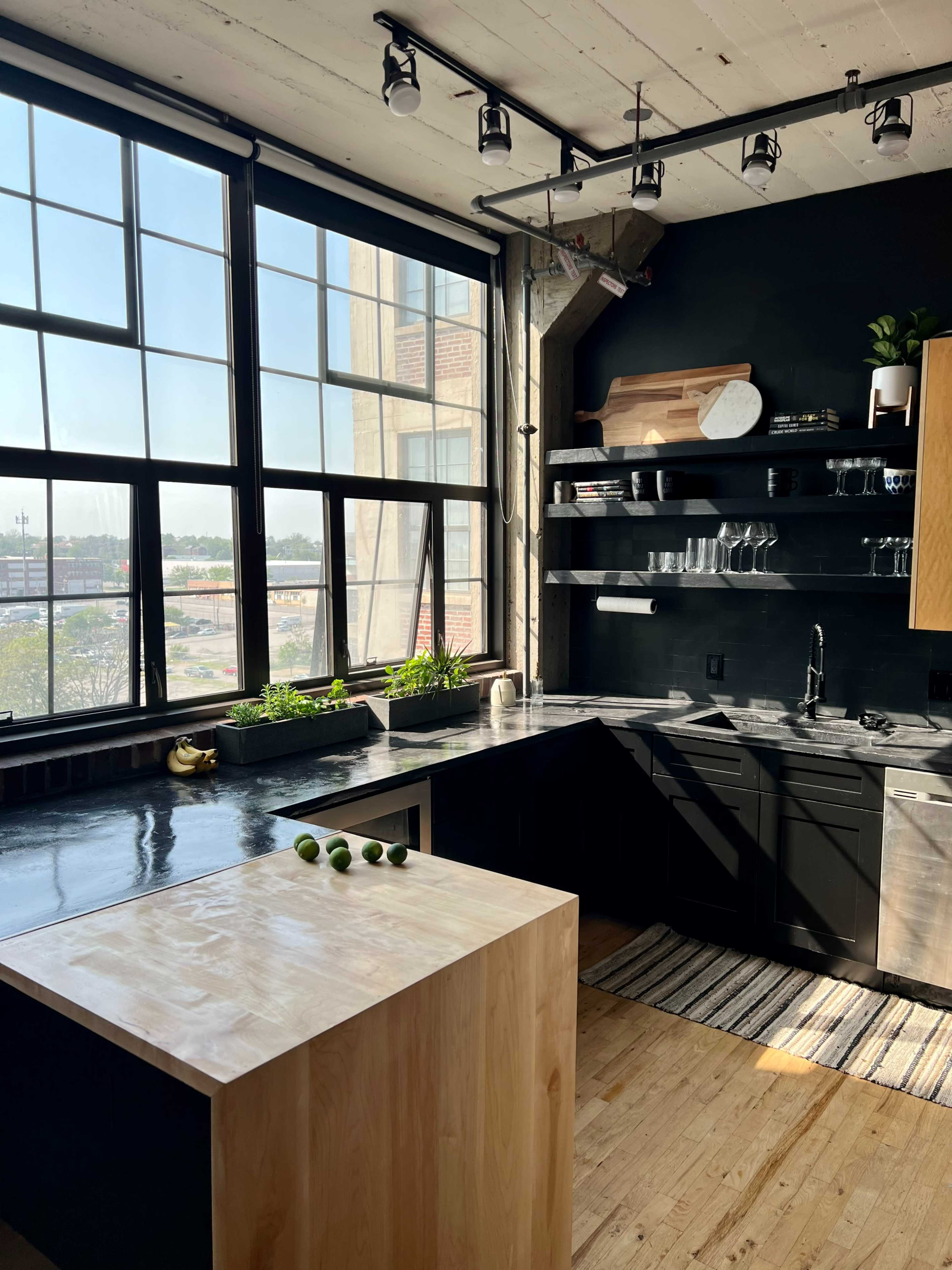 A modern kitchen featuring a large window, dark cabinetry, a minimalist countertop, and a wooden island with three limes placed on it.