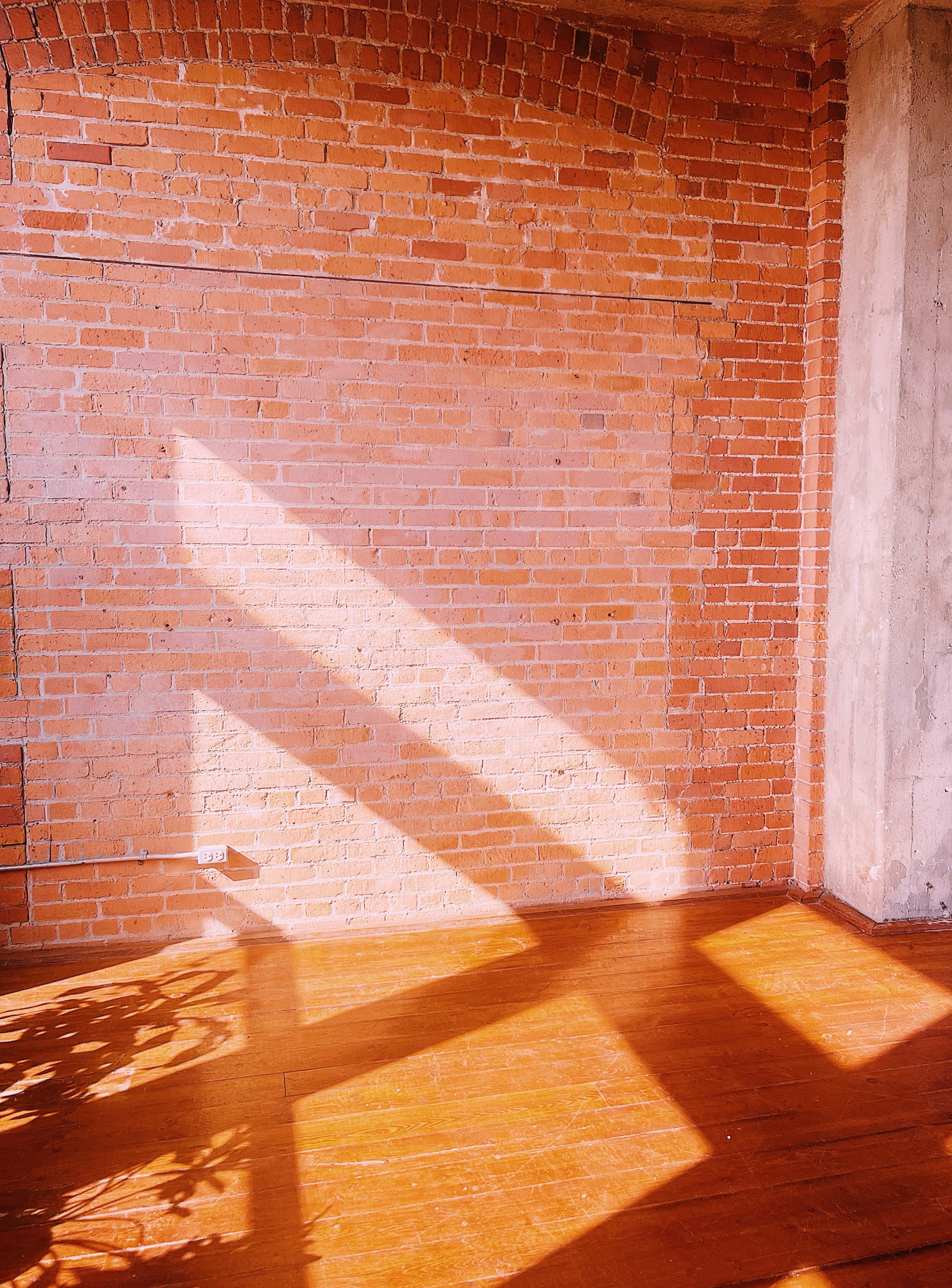 Sunlight casts long shadows across a wooden floor and exposed brick wall in an empty room.