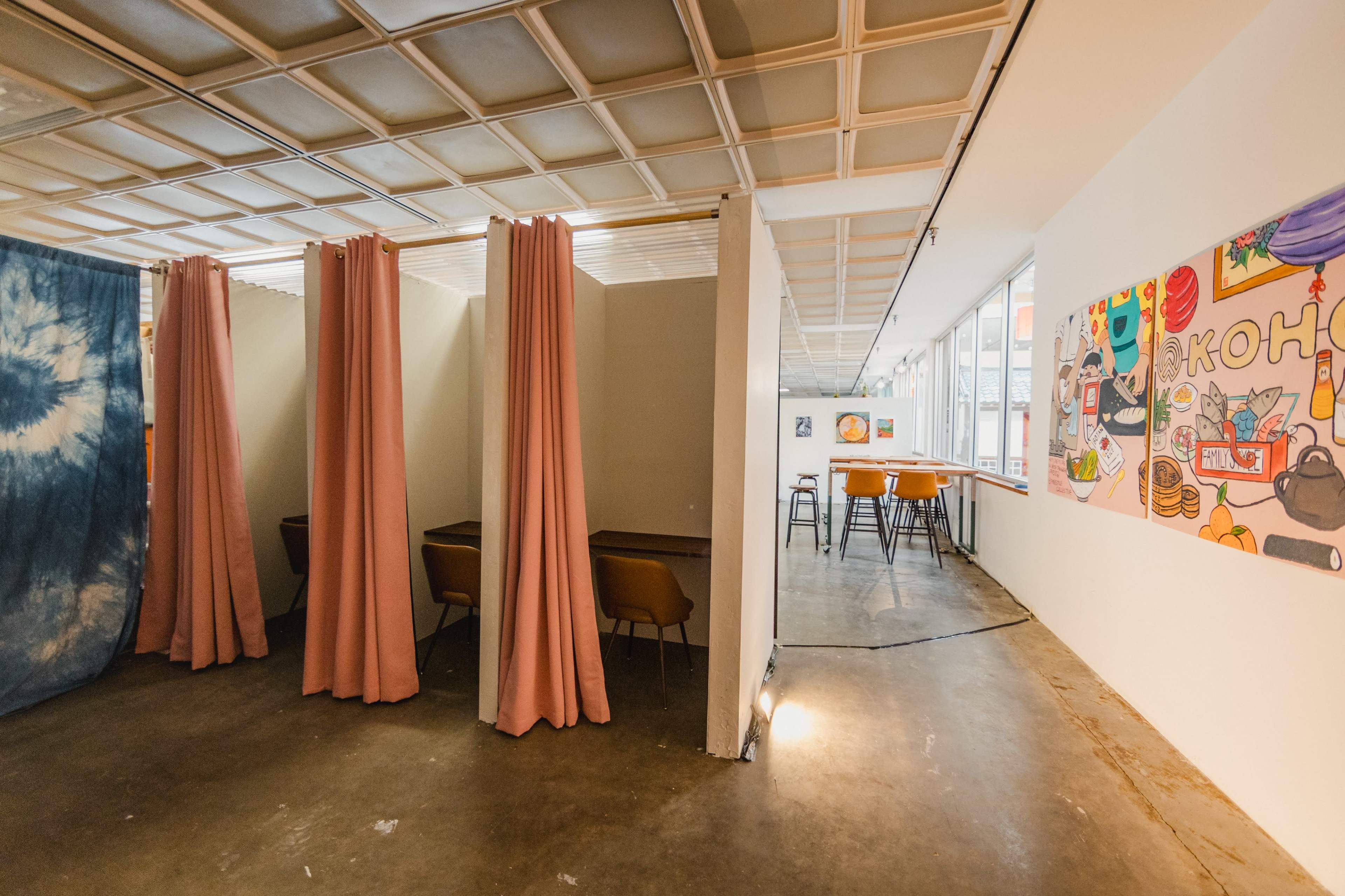 The image shows a modern workspace with booth-style seating separated by fabric curtains, alongside a brightly decorated wall featuring colorful artwork.