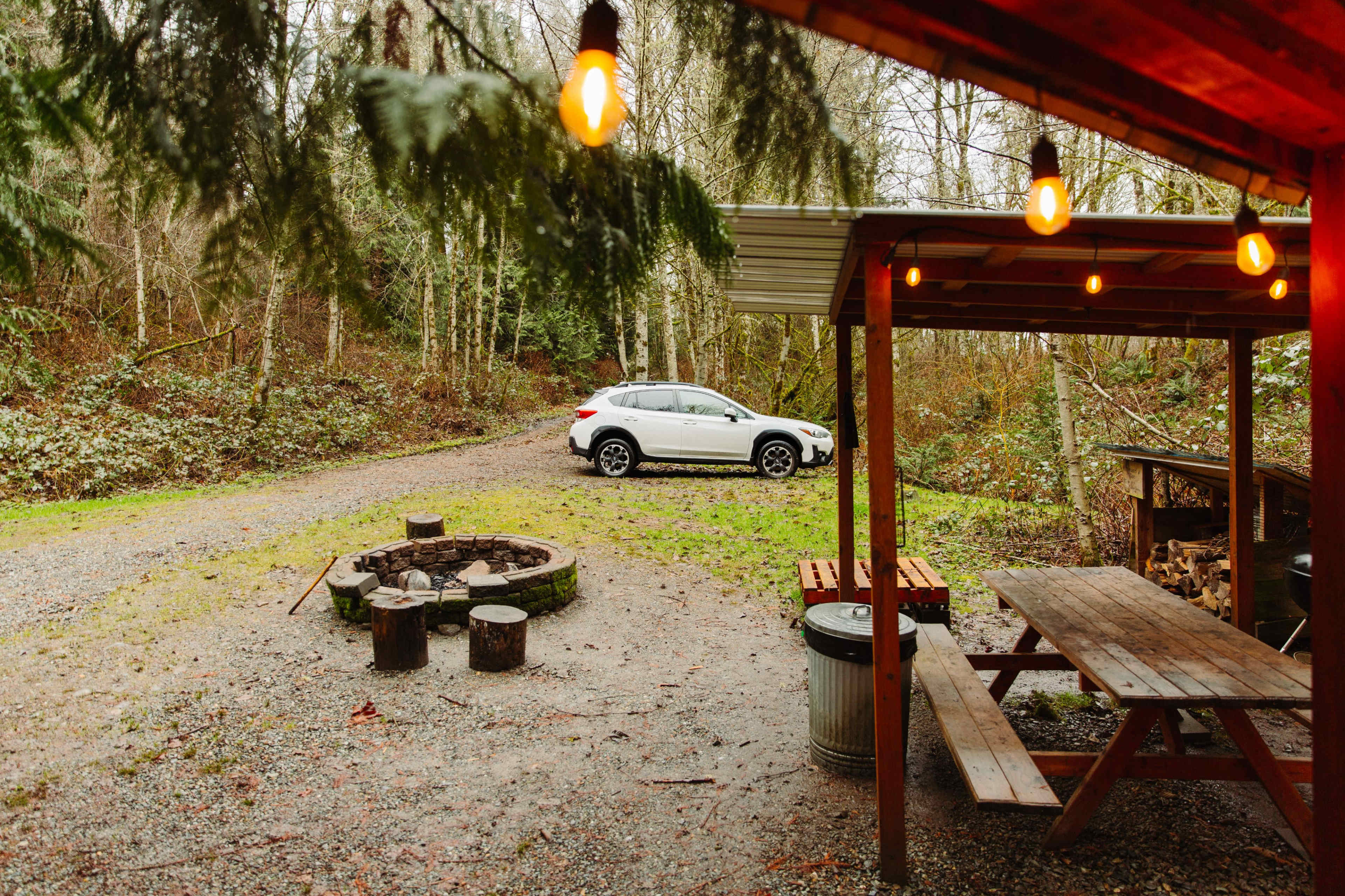 A gray SUV is parked on a gravel path next to a fire pit surrounded by wooden seating and a picnic table under a covered area.