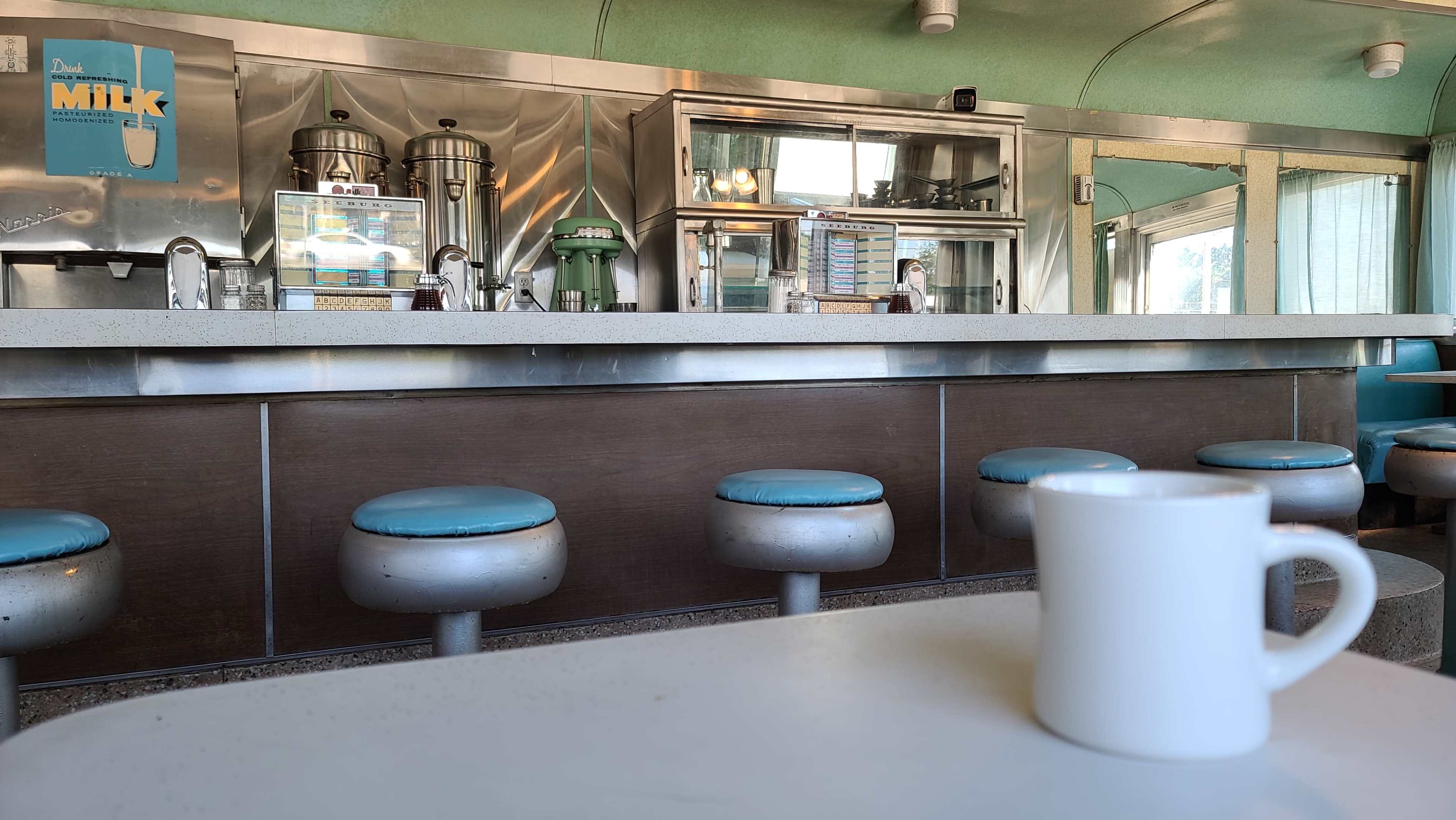 A diner interior with metallic countertops, teal stools, and a white coffee cup in the foreground.