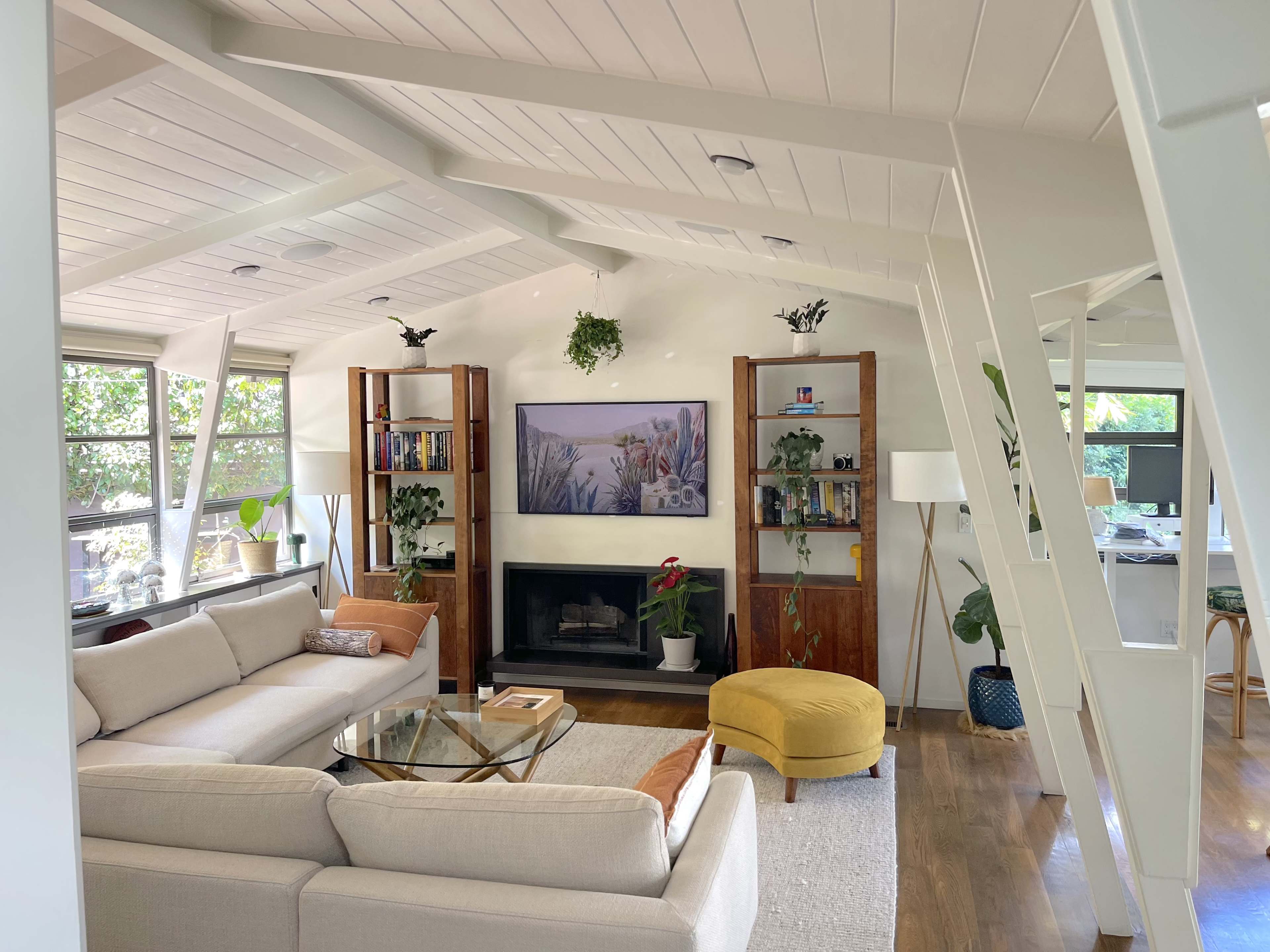 A modern living room featuring a beige sectional sofa, a glass coffee table, a fireplace with a framed artwork above it, and wooden bookshelves on either side.