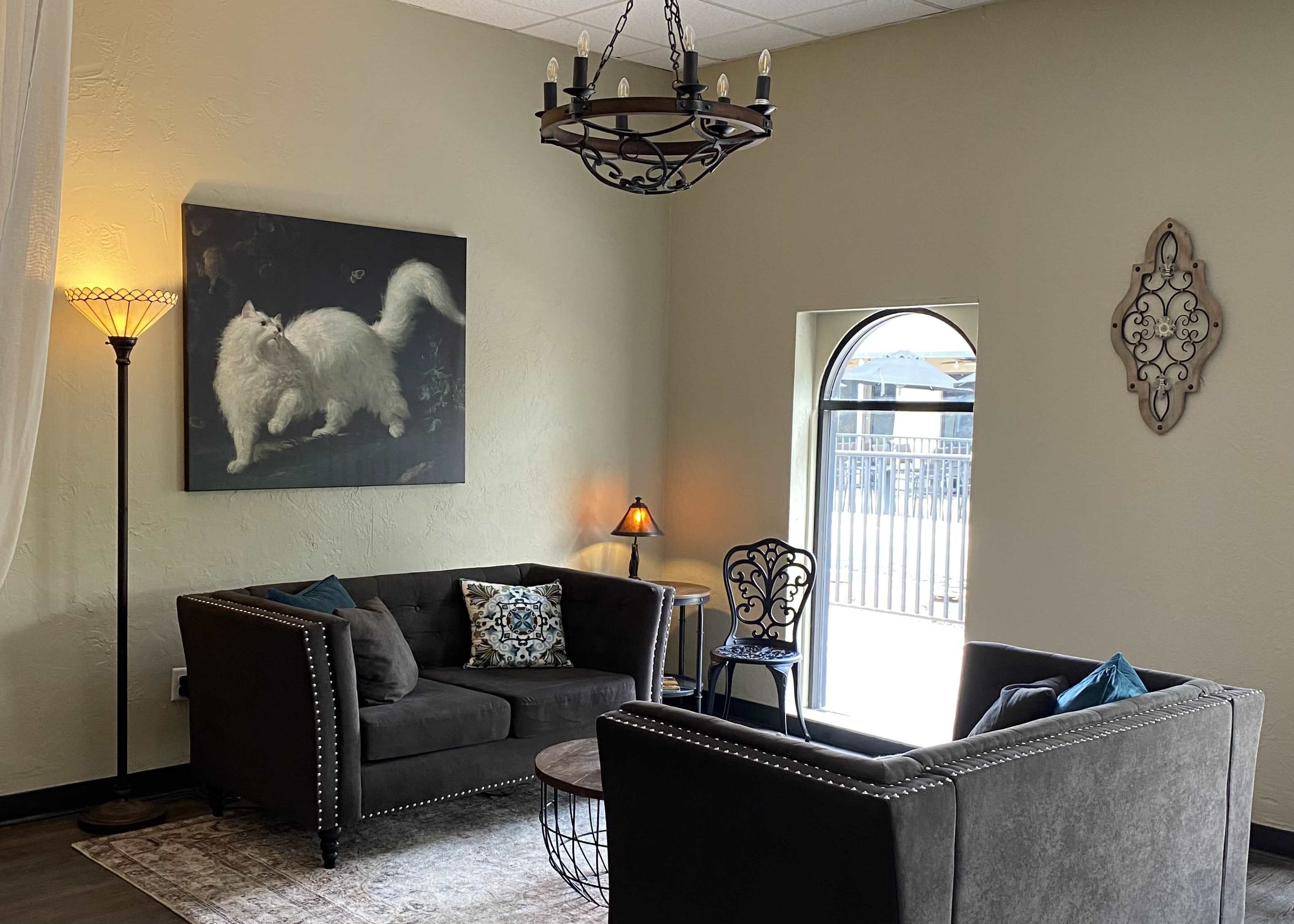 A living room with two dark sofas, a coffee table, and artwork of a white cat on the wall, illuminated by a large chandelier and a floor lamp.