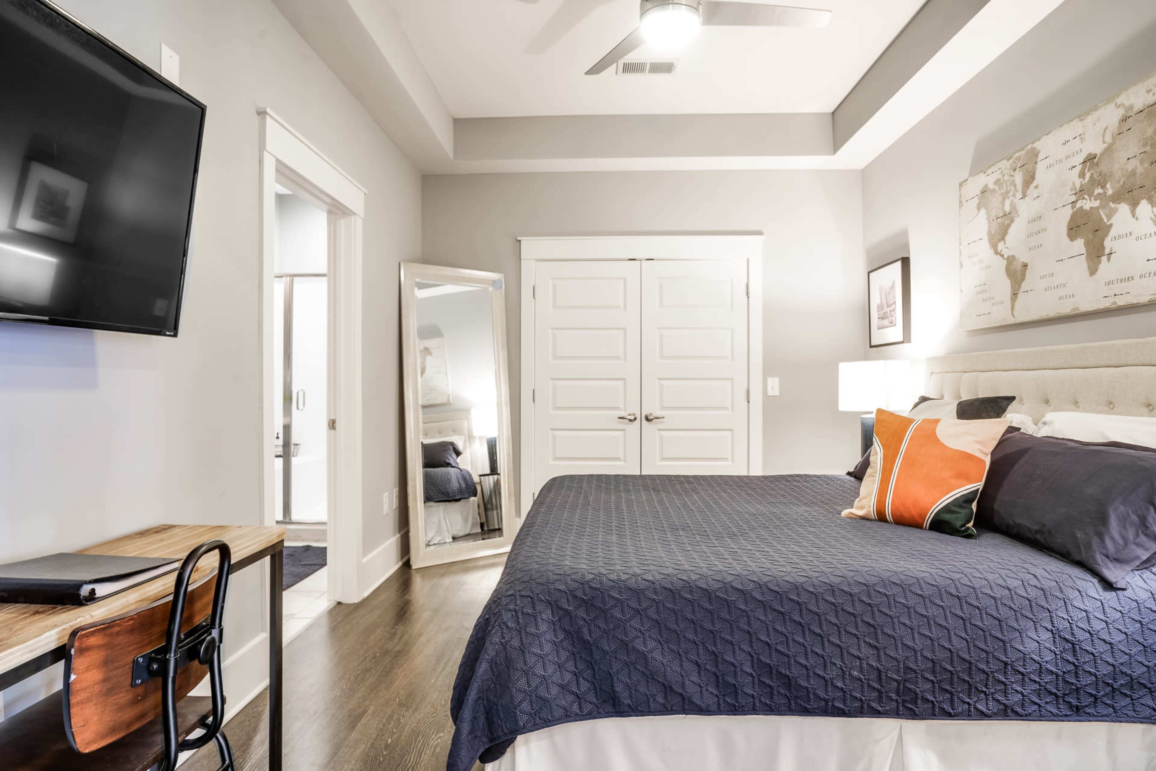 The image shows a modern bedroom with a neatly made bed, a wall-mounted TV, and a large mirror reflecting the room's interior.