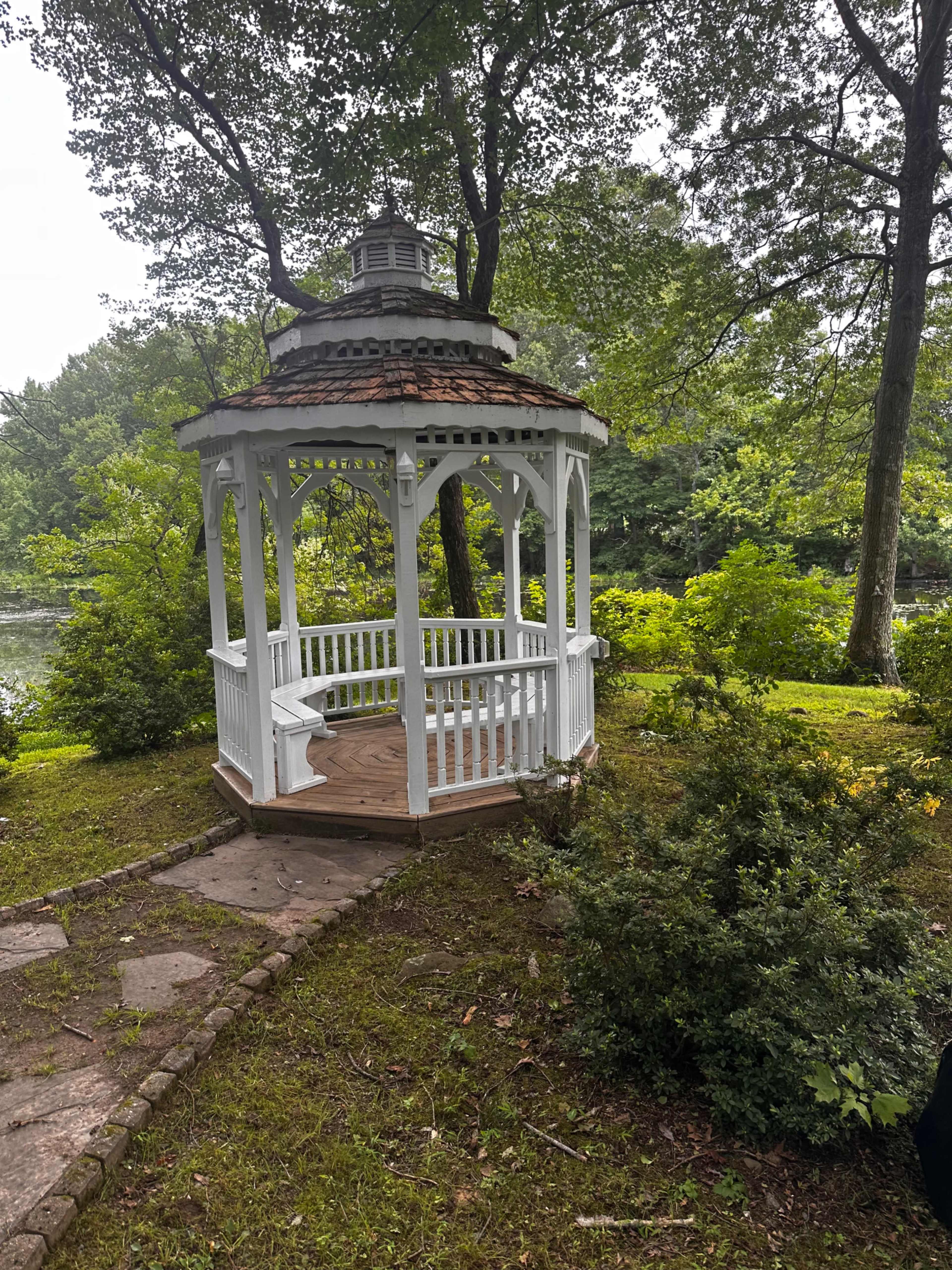 A white gazebo with a conical roof is situated on a grassy area surrounded by trees and shrubs near a body of water.