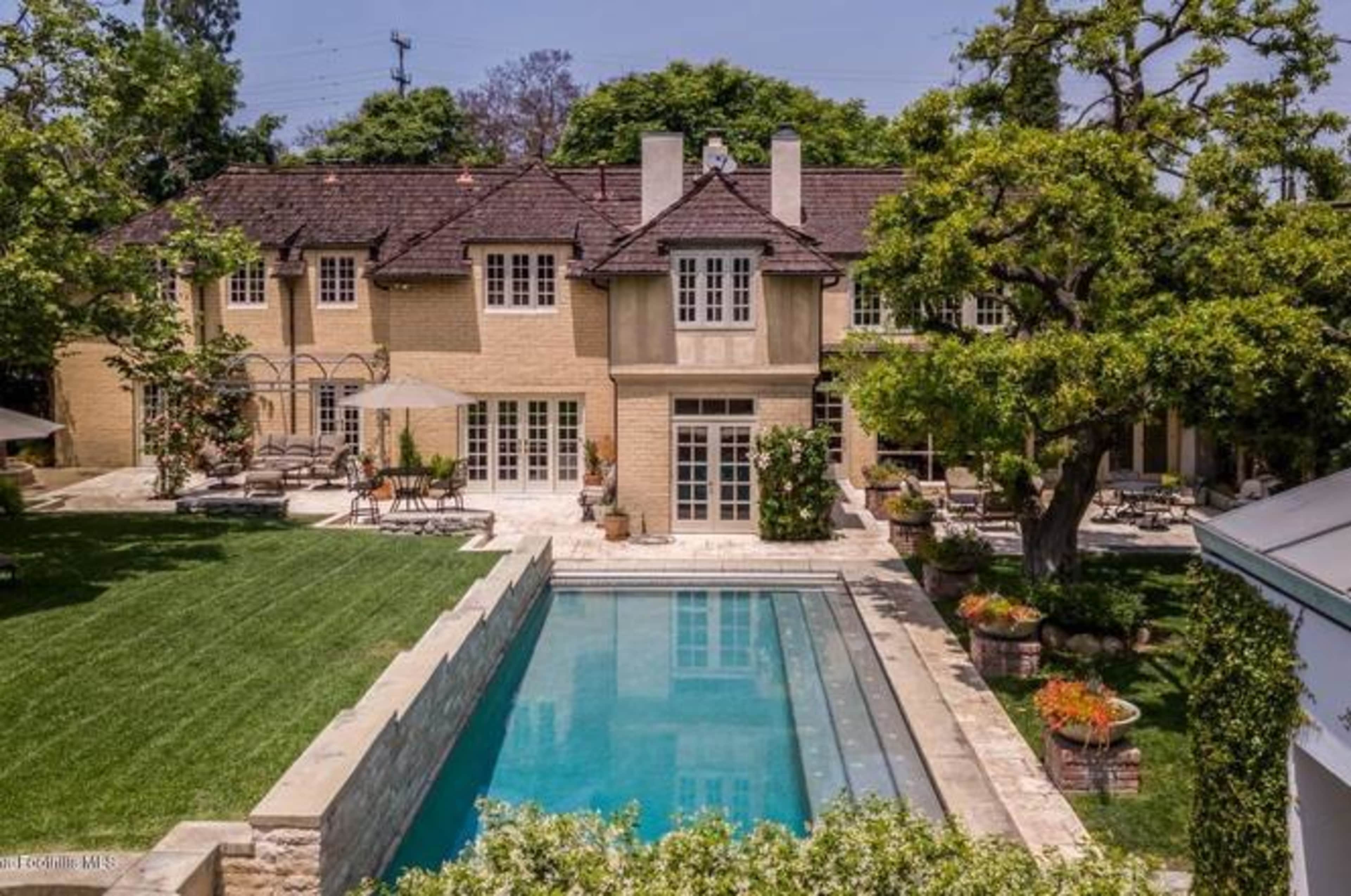 The image shows a large, multi-story house with a pool and landscaped lawn, surrounded by trees and lounge areas.