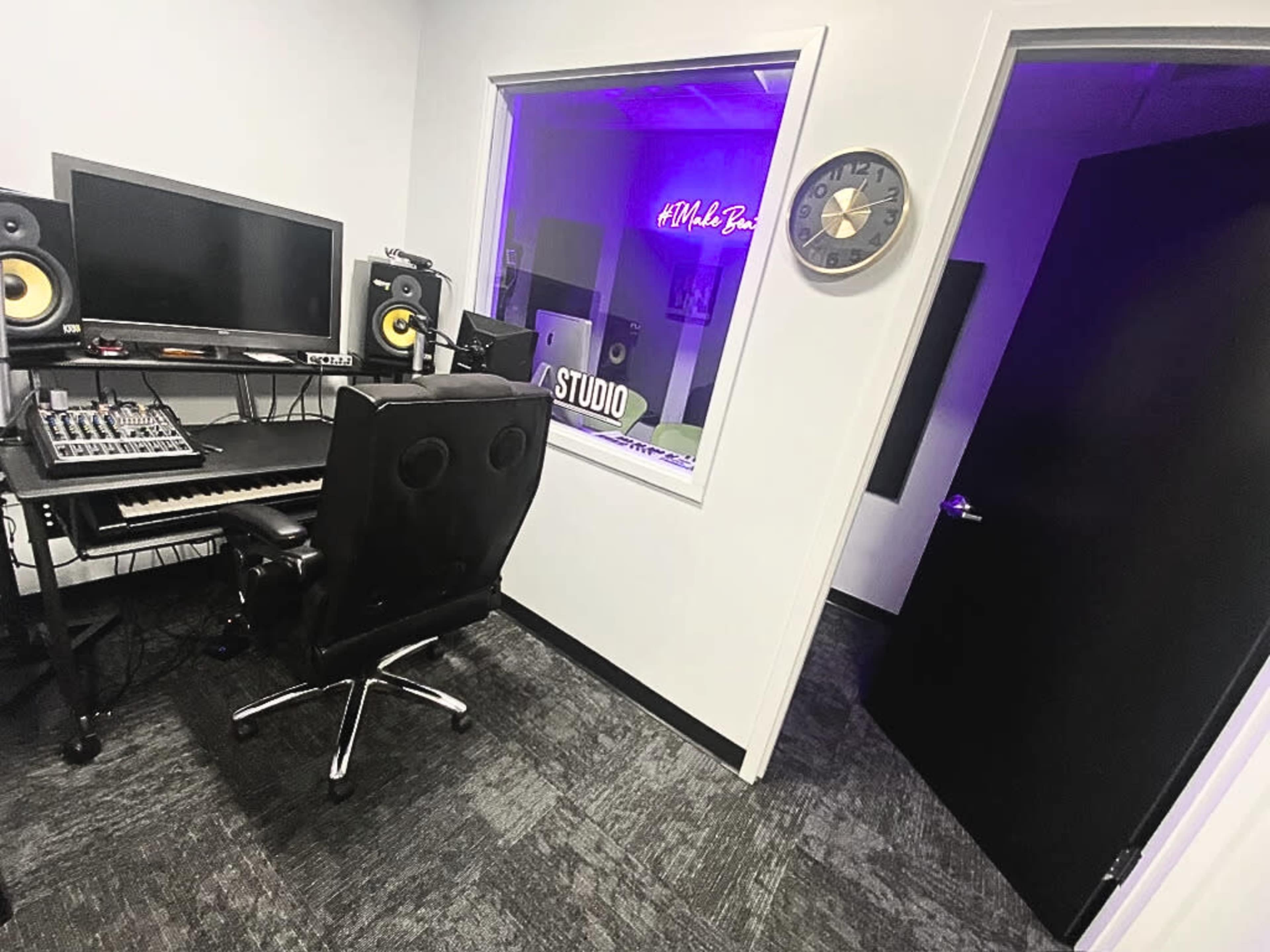 A music studio with a desk equipped with audio equipment, a computer monitor, and speakers, beside a glass-walled room containing a sign that reads "Hustle Boss."