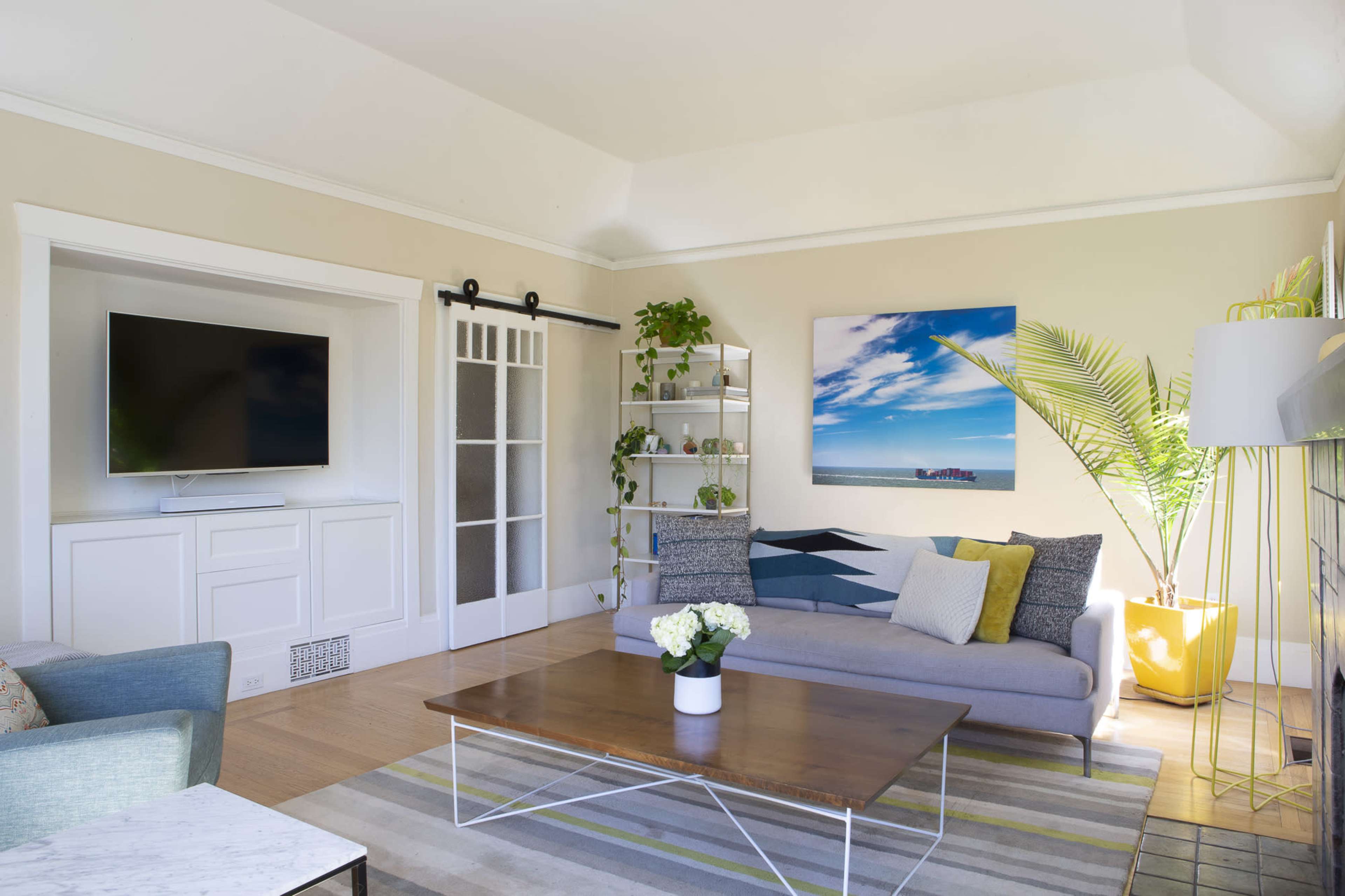 A bright living room featuring a gray sofa, a wooden coffee table, and a large television mounted on the wall, with decorative plants and artwork enhancing the space.