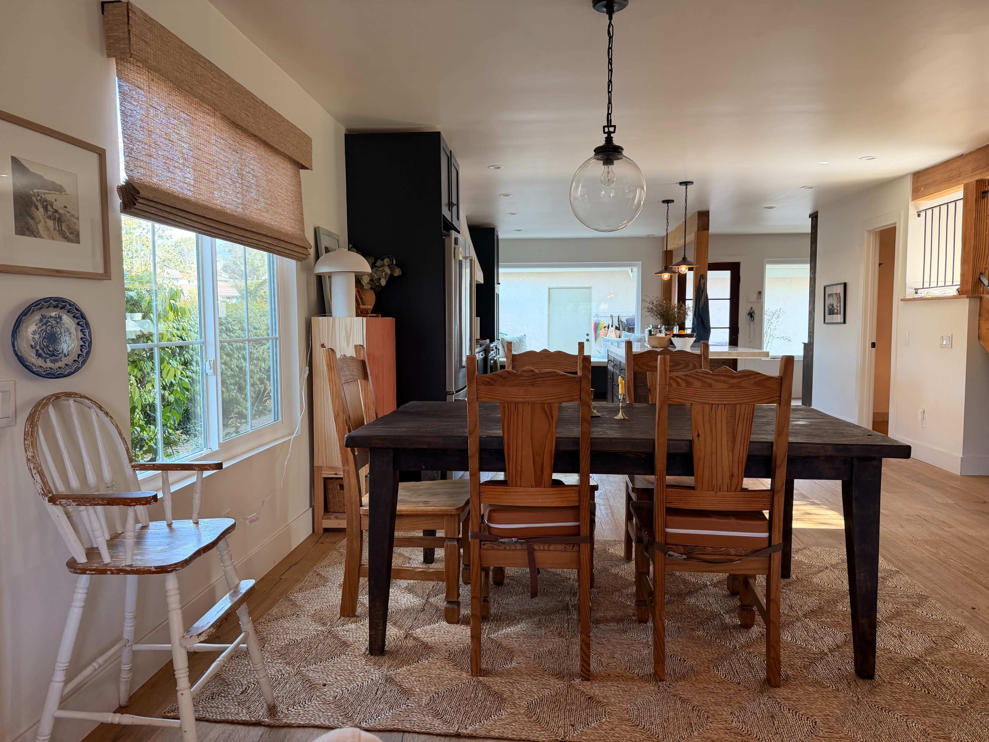 The image shows a dining area with a wooden table surrounded by chairs, a high chair in the corner, and large windows allowing natural light to illuminate the space.