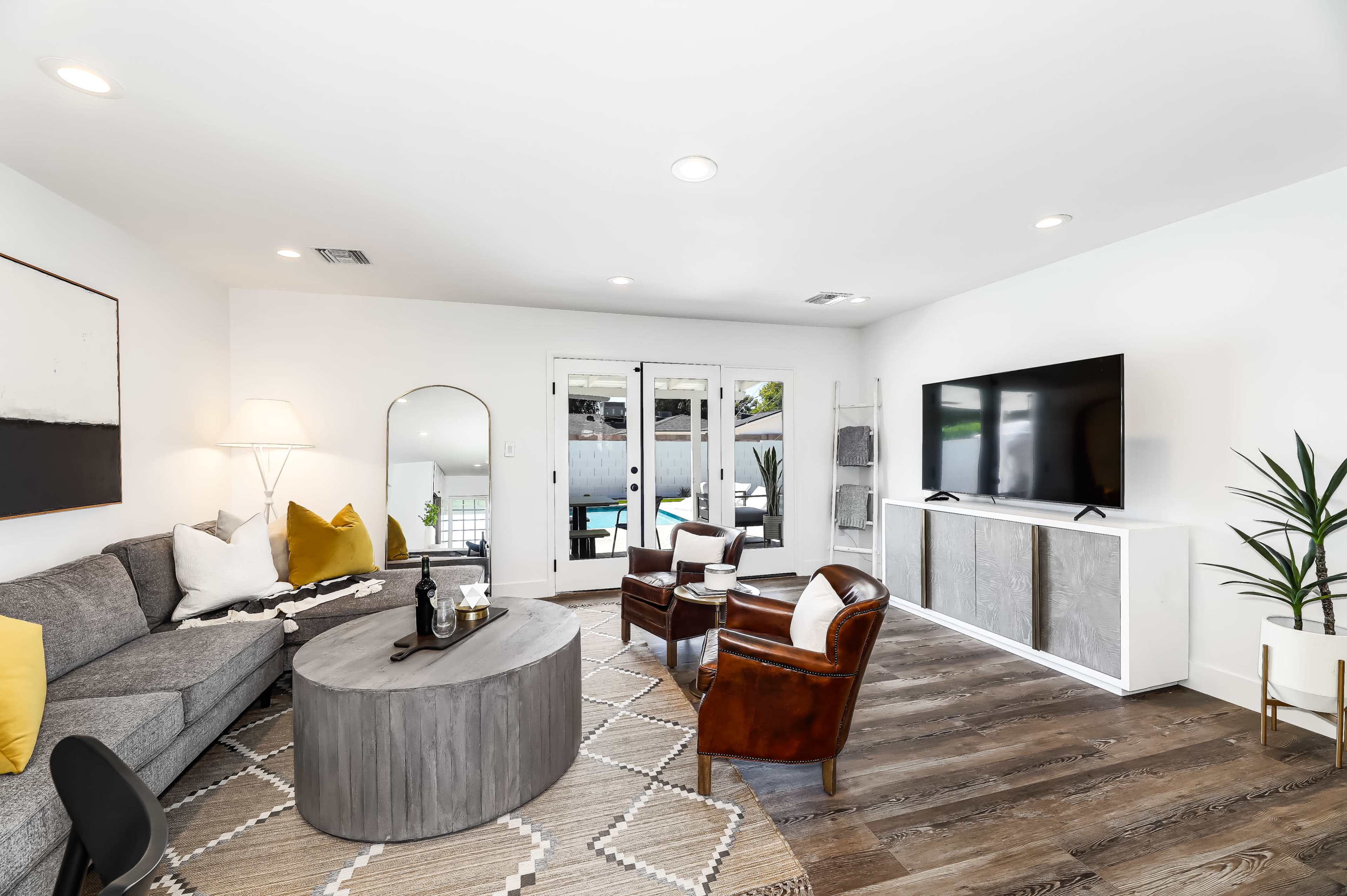 The living room features a gray sectional sofa, a circular coffee table, and a large television, with a view of a pool visible through glass doors.