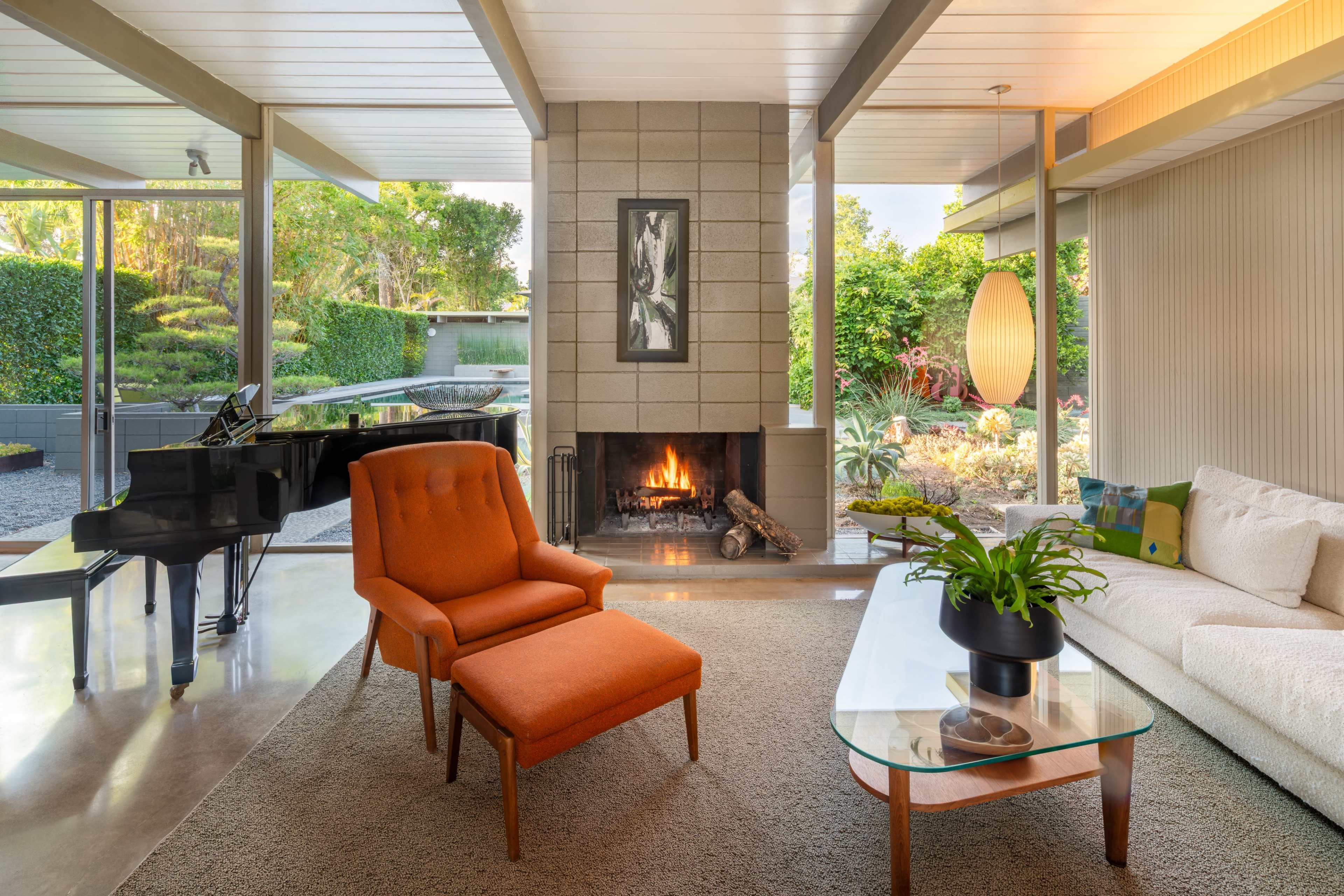 A modern living room features a fireplace, an orange armchair, a glass coffee table, and large windows overlooking a landscaped garden.