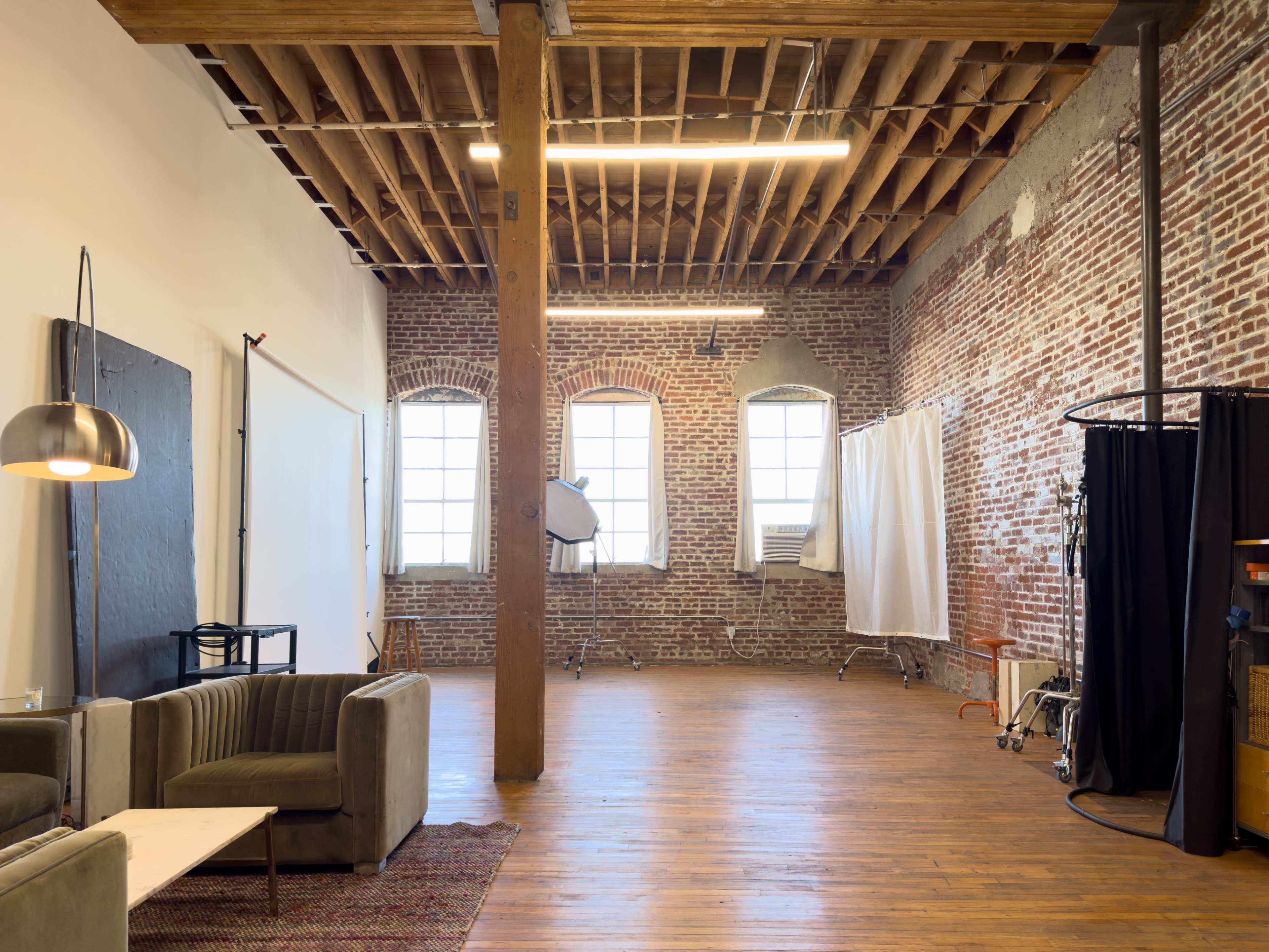 The image shows a spacious studio with exposed brick walls, hardwood flooring, and large windows, featuring a seating area and various photo equipment.