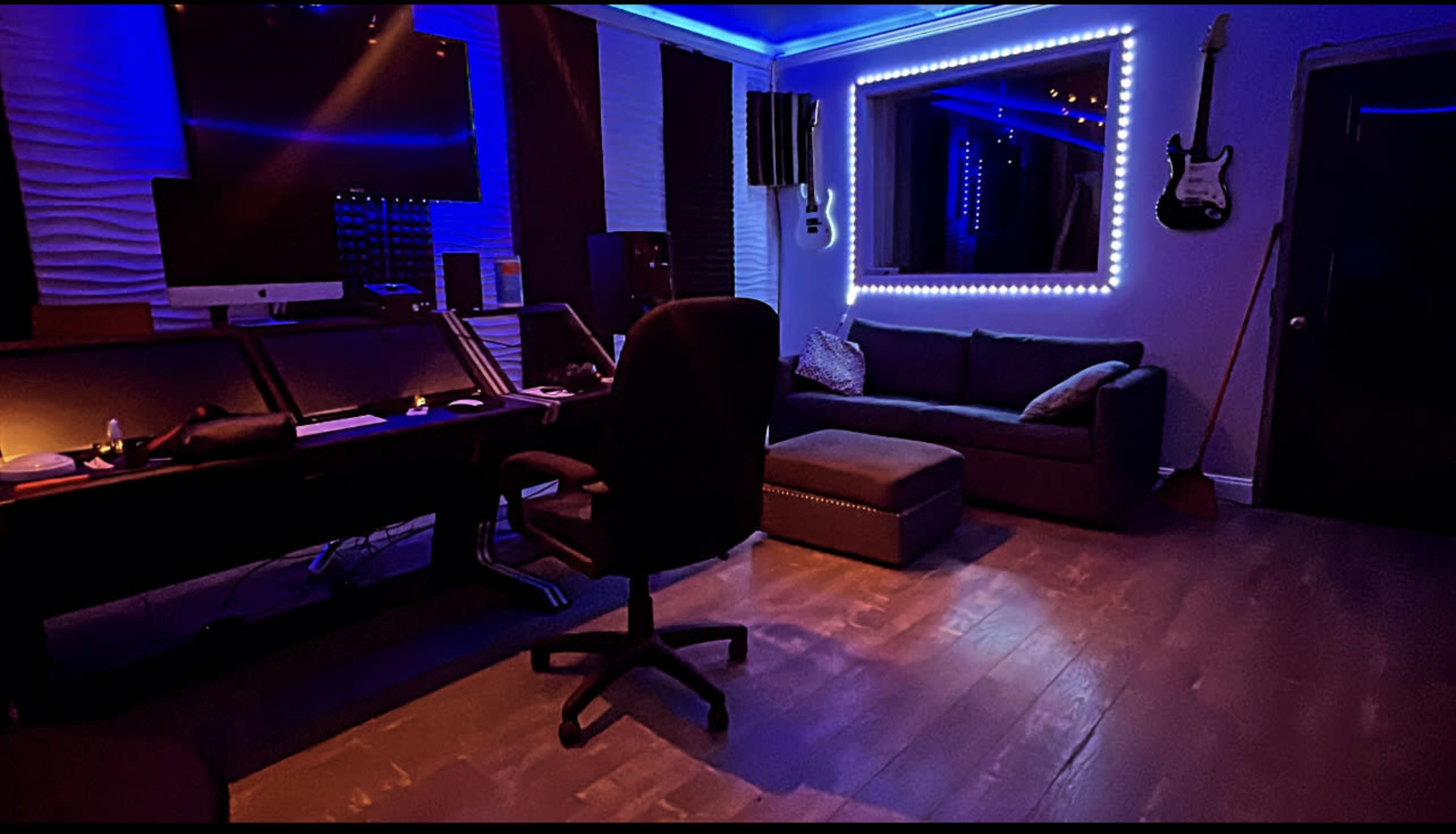 A music studio featuring a desk with computer equipment, a black swivel chair, a couch, and illuminated wall decorations.