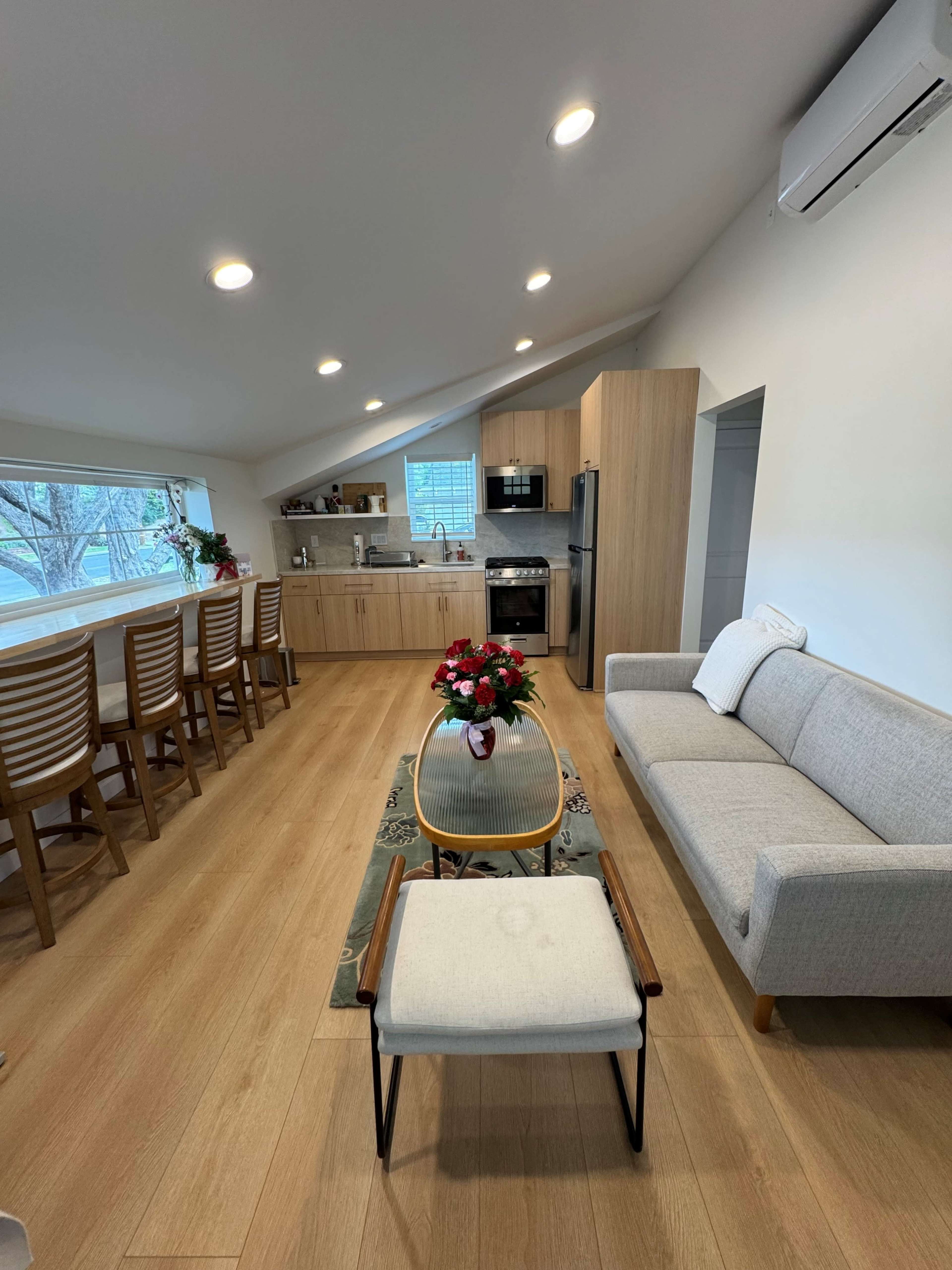 The image shows a modern kitchen and living area with light wood flooring, a gray sofa, and a dining space with bar stools.