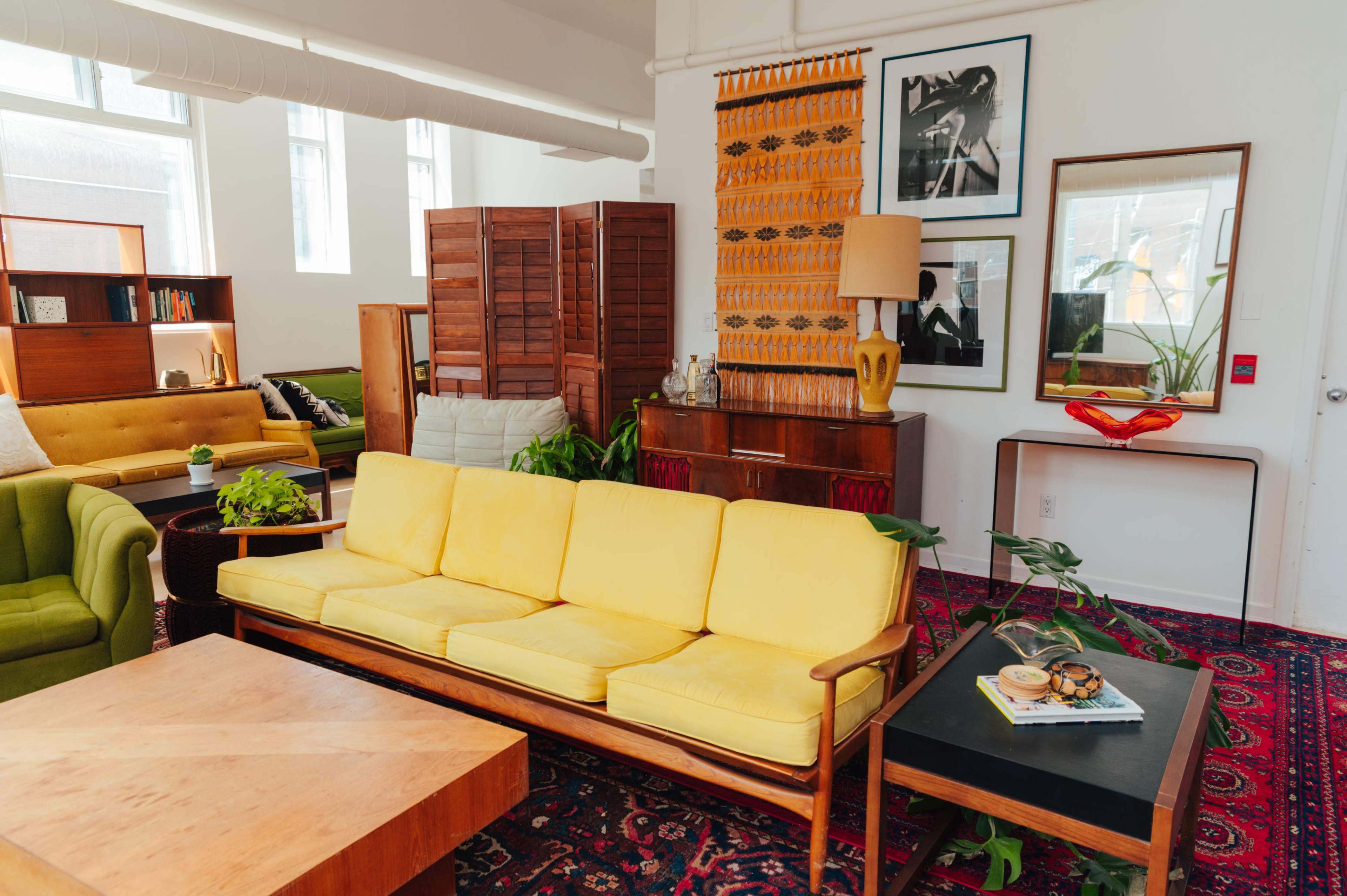 A brightly lit living room featuring a yellow couch, a wooden coffee table, and various decorative elements like plants and artwork.