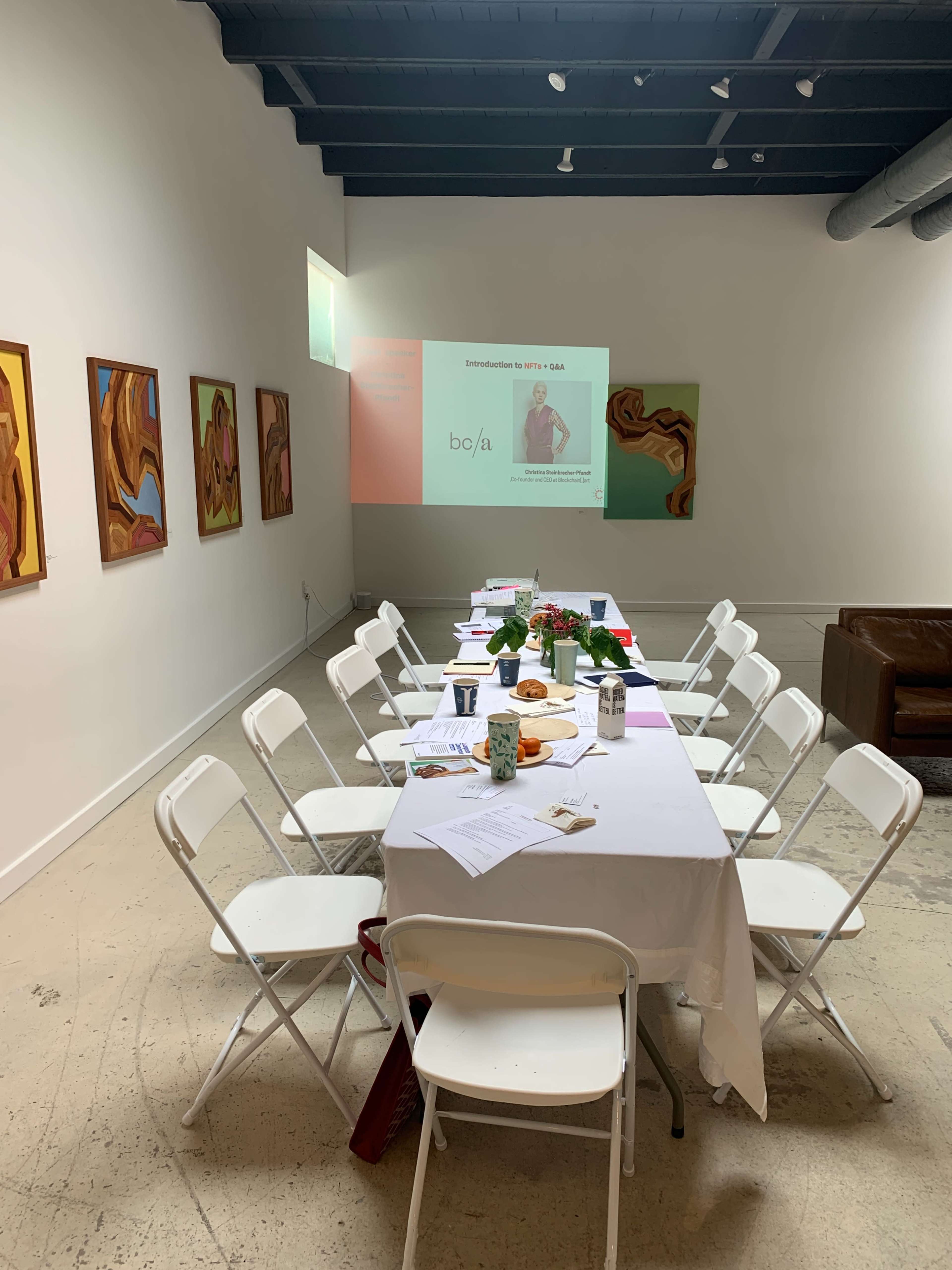 A long table with white chairs is set up in a bright room, featuring a presentation screen and several artworks on the walls.