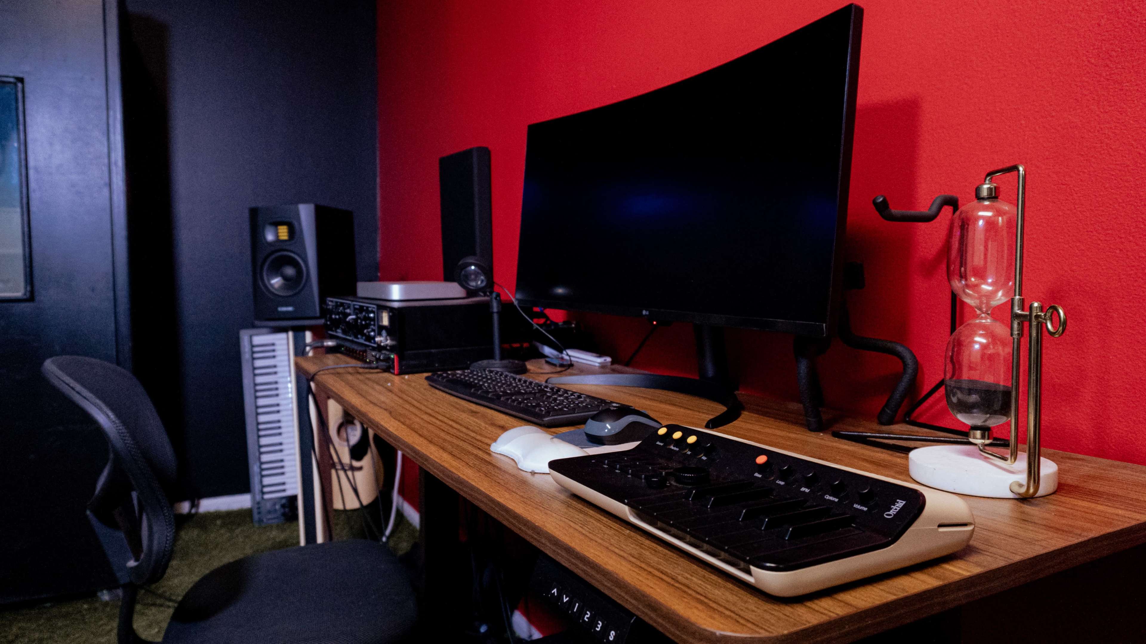 A modern desk setup features an ultrawide monitor, an audio interface, studio speakers, and a musical keyboard against a red wall.