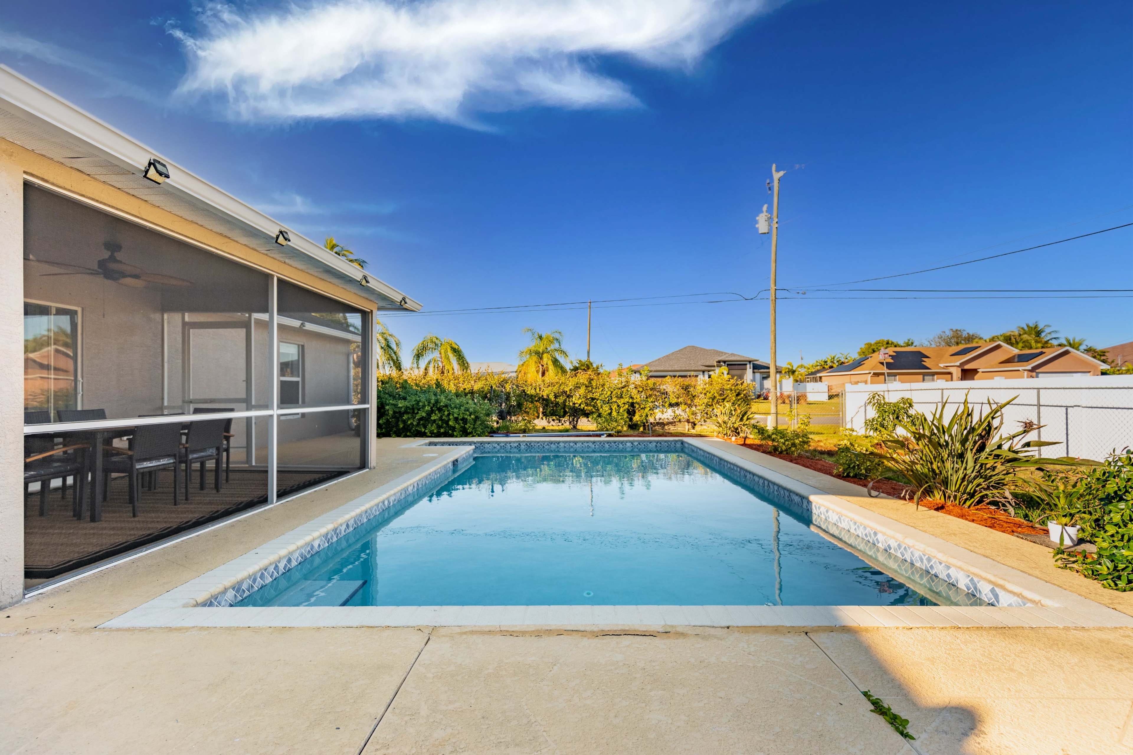 A clear swimming pool sits in an outdoor area surrounded by greenery and a fence under a bright blue sky.
