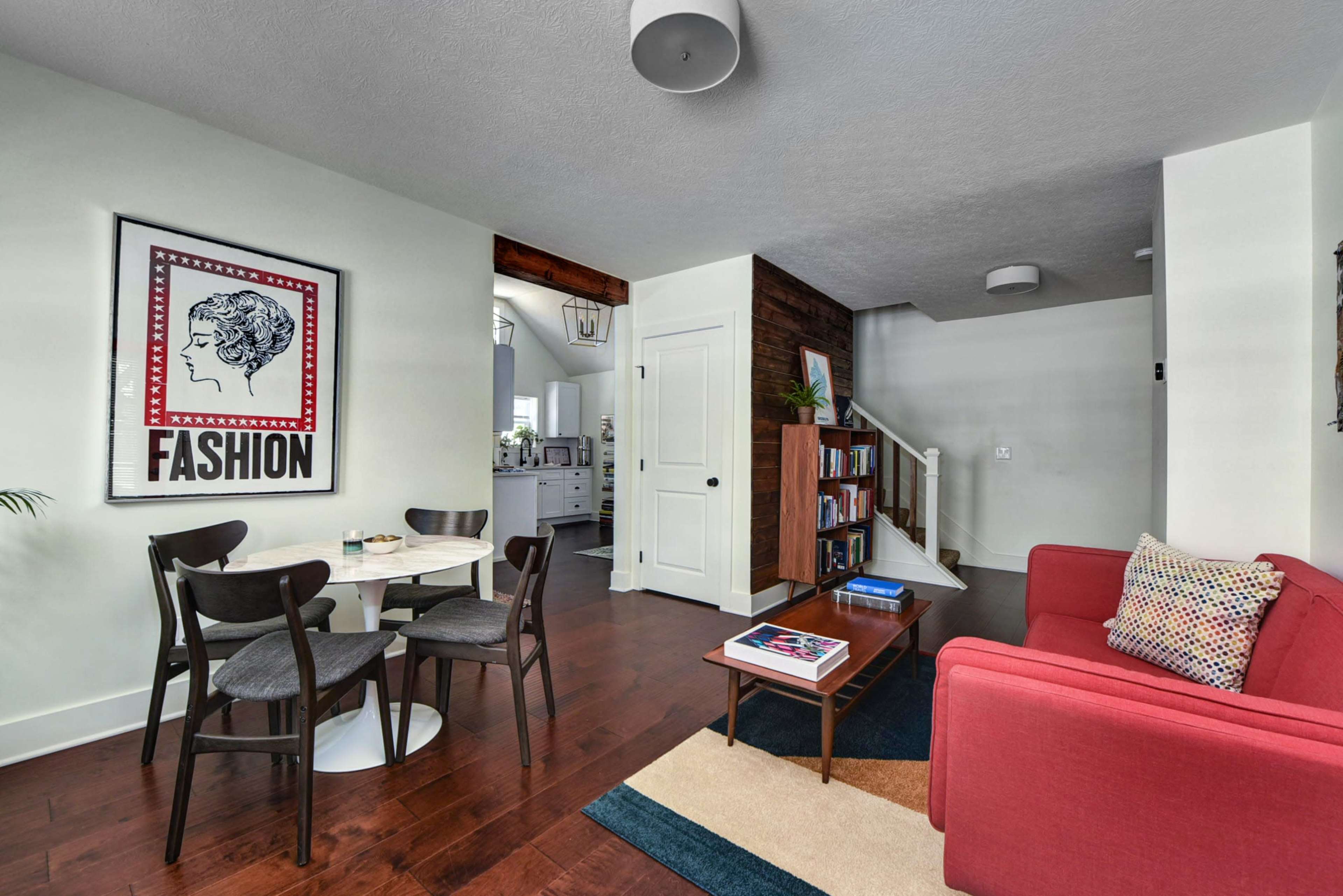 The interior features a dining area with a round table and four chairs, a red couch, and access to an adjacent staircase and living space.