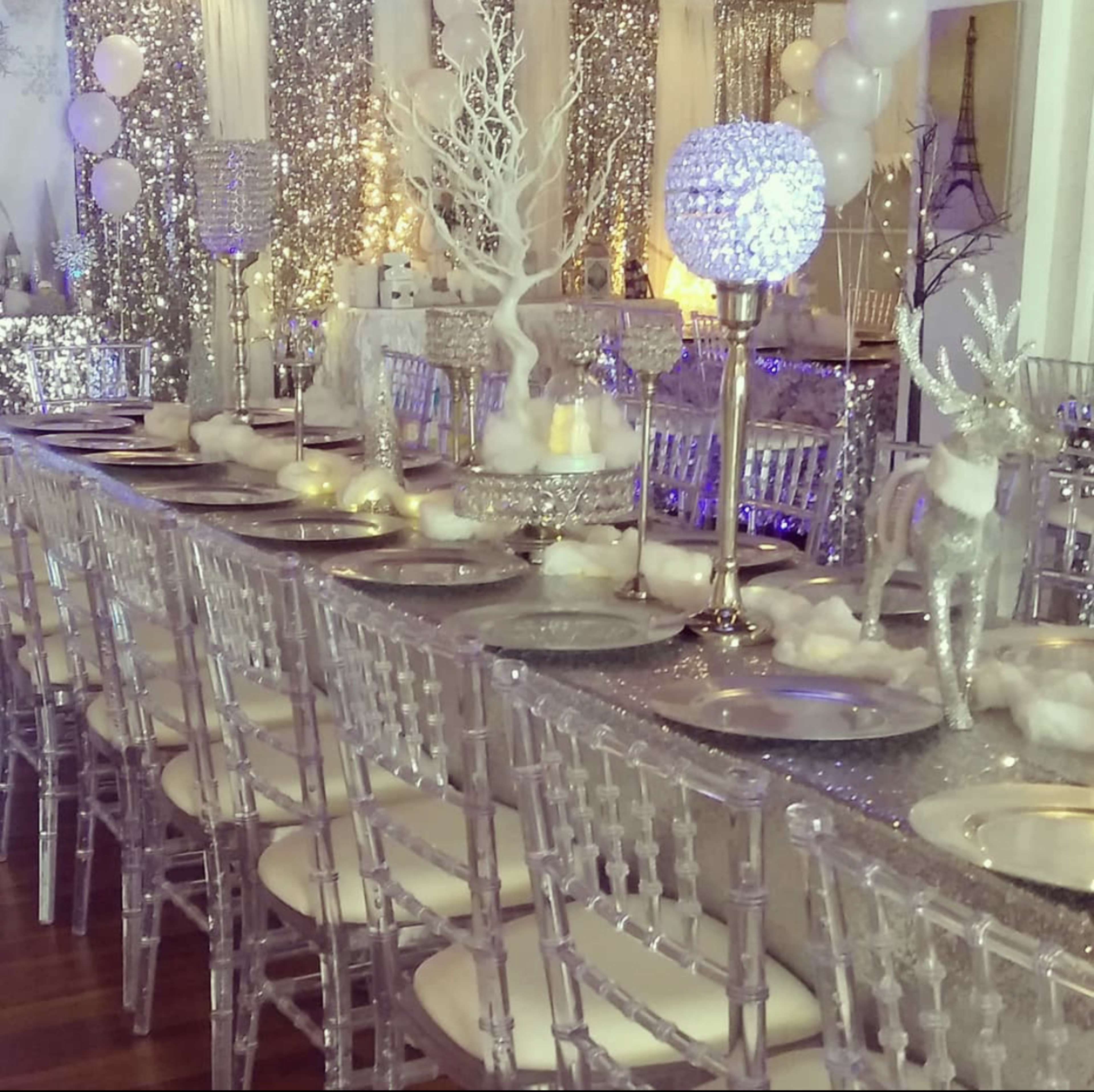 A long dining table is set with silver decorations, clear chairs, and winter-themed centerpieces, including reindeer and lit orbs, in a sparkling event space.