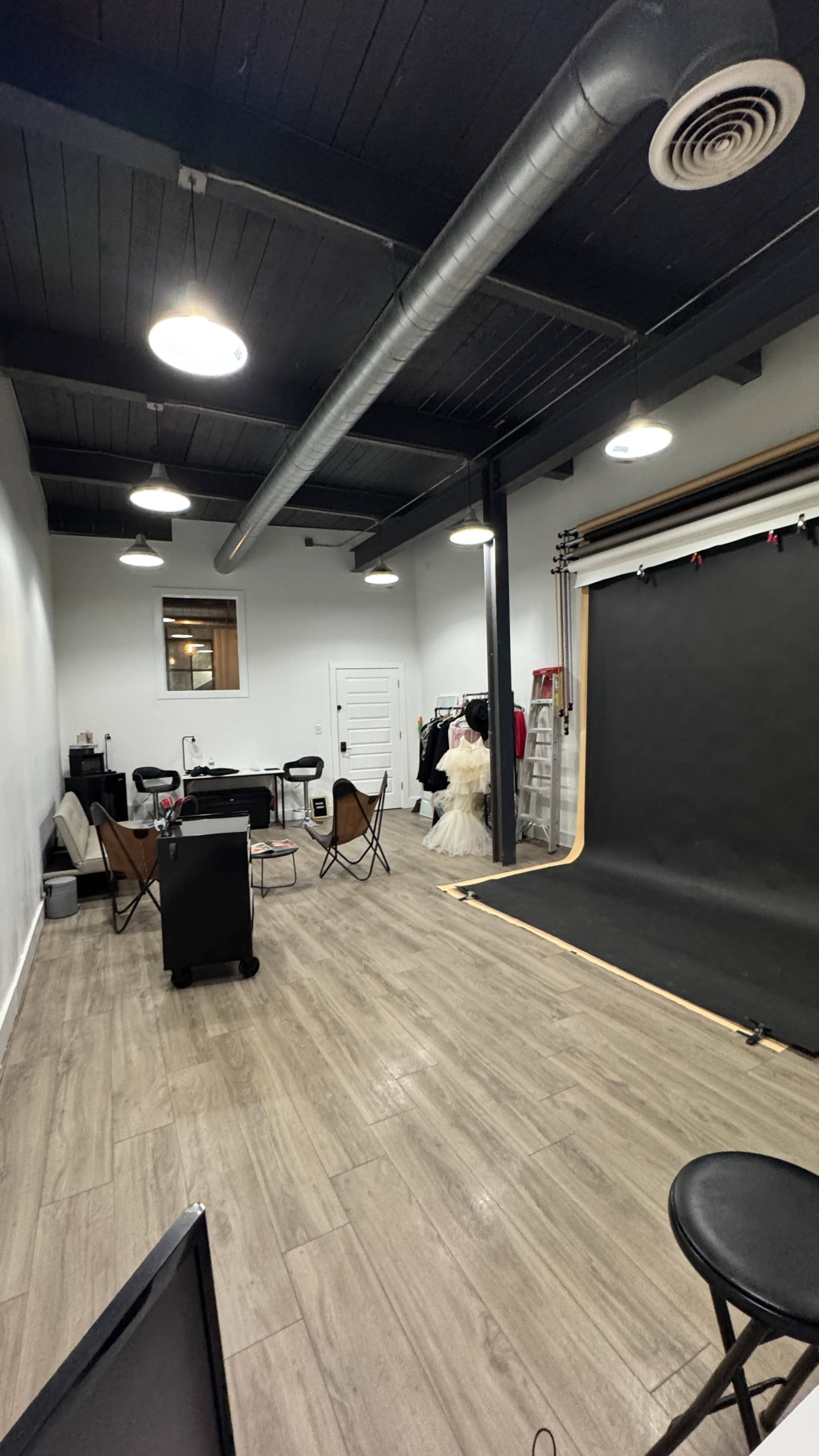 A spacious studio with wooden flooring features a photo backdrop, chairs, a table, and a dress hanging on a rack.