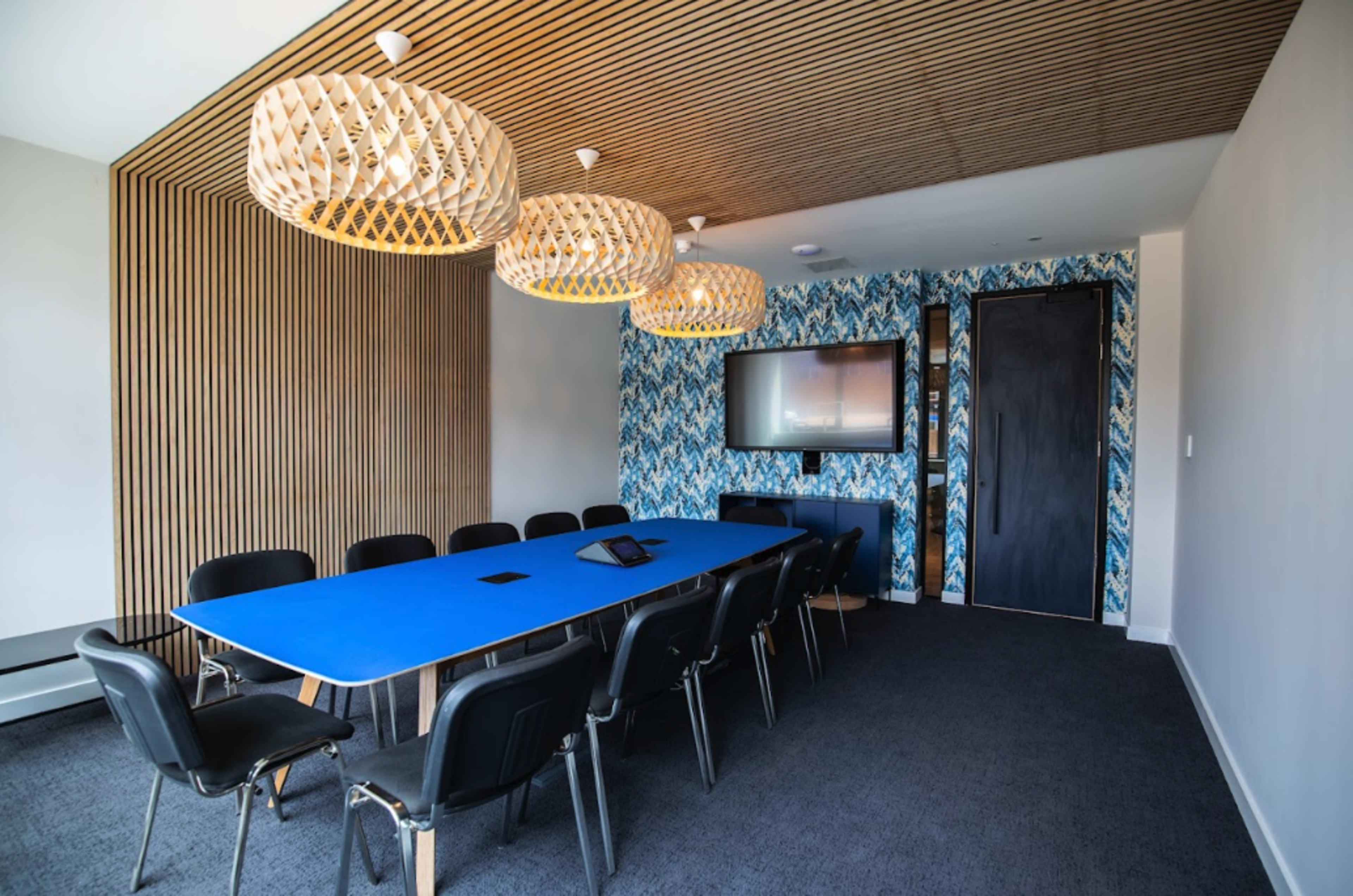 A modern conference room features a long blue table surrounded by black chairs, with patterned wallpaper and pendant lights above.