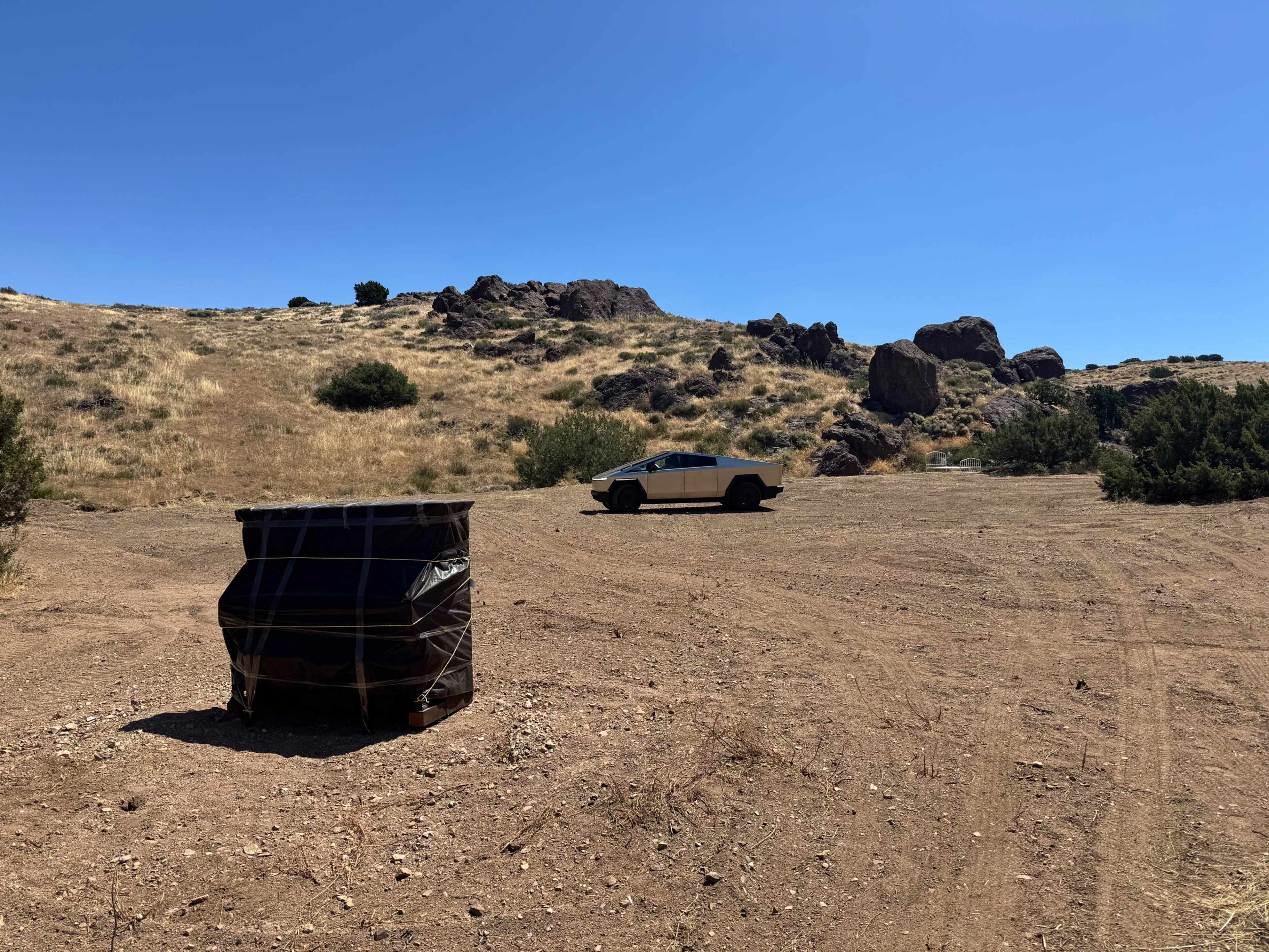 A wrapped object is situated on a dirt patch with sparse vegetation, while a futuristic vehicle is parked in the background against a rocky hillside under a clear blue sky.