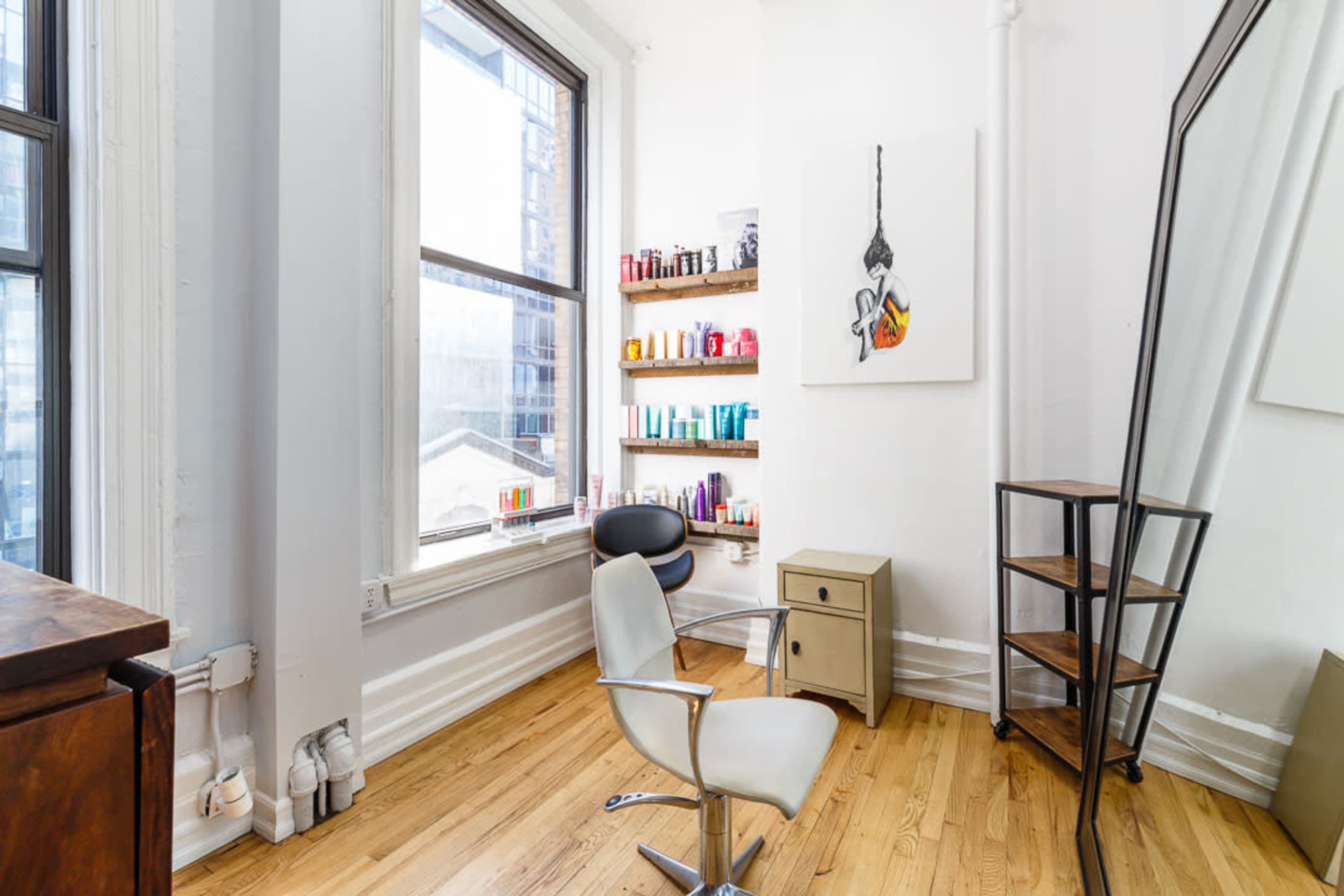 The image shows a minimalist hair salon interior featuring a chair, a large mirror, a shelf with various hair products, and wooden flooring.