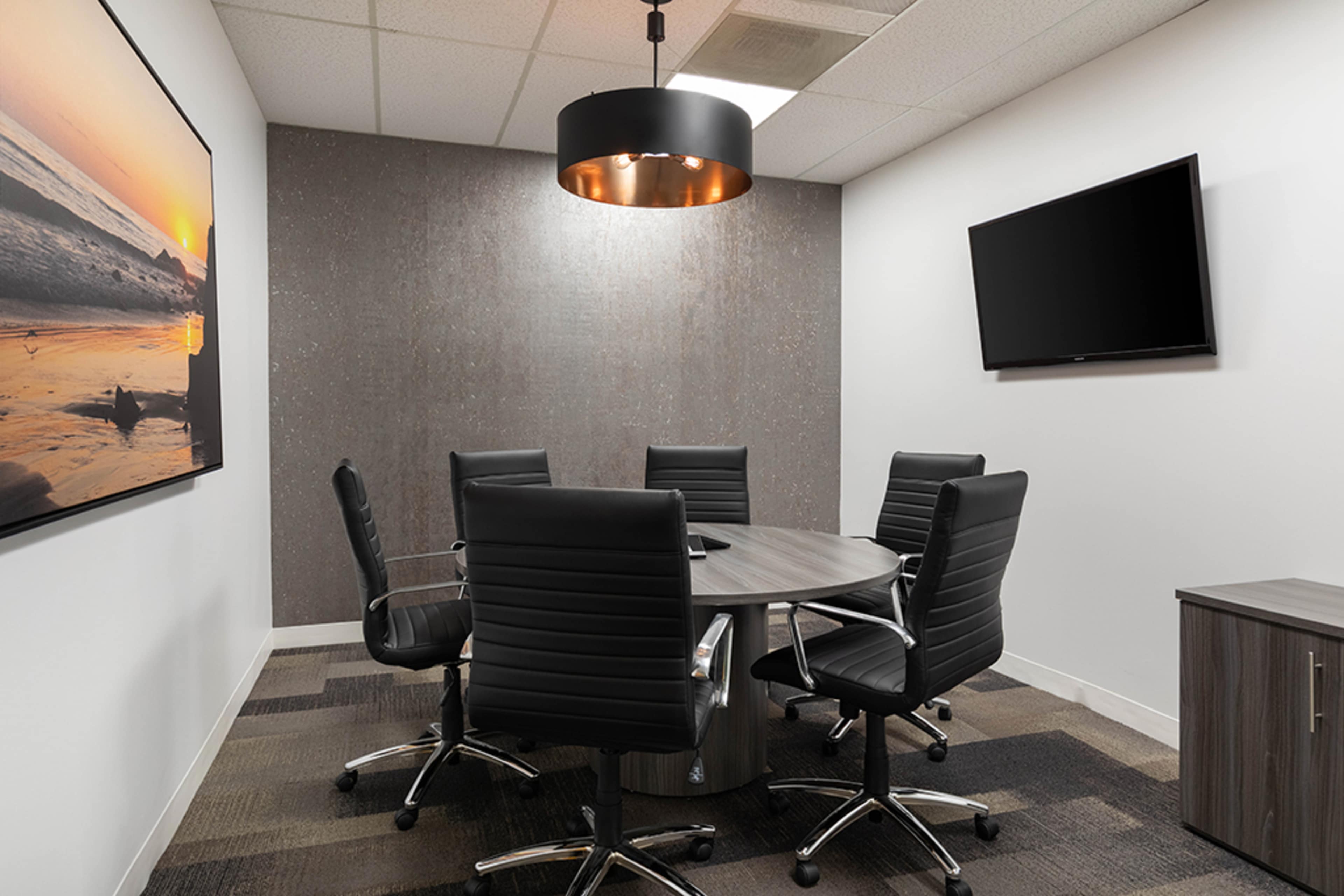 A conference room features a round table surrounded by five black rolling chairs, with an overhead light and a wall-mounted television.