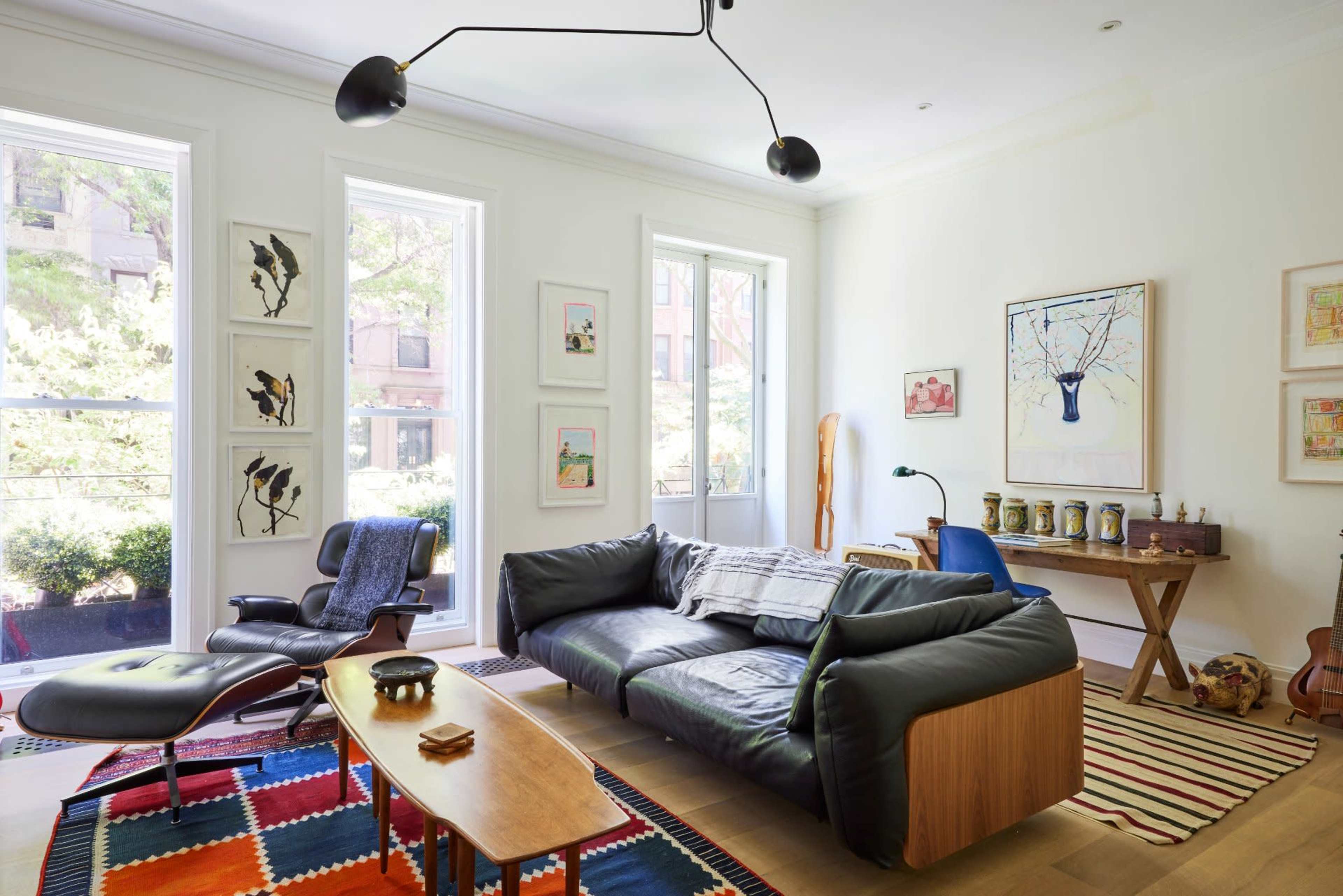 The image shows a modern living room with a black leather sofa, a wooden coffee table, an armchair, and large windows letting in natural light, adorned with various artworks on the walls.