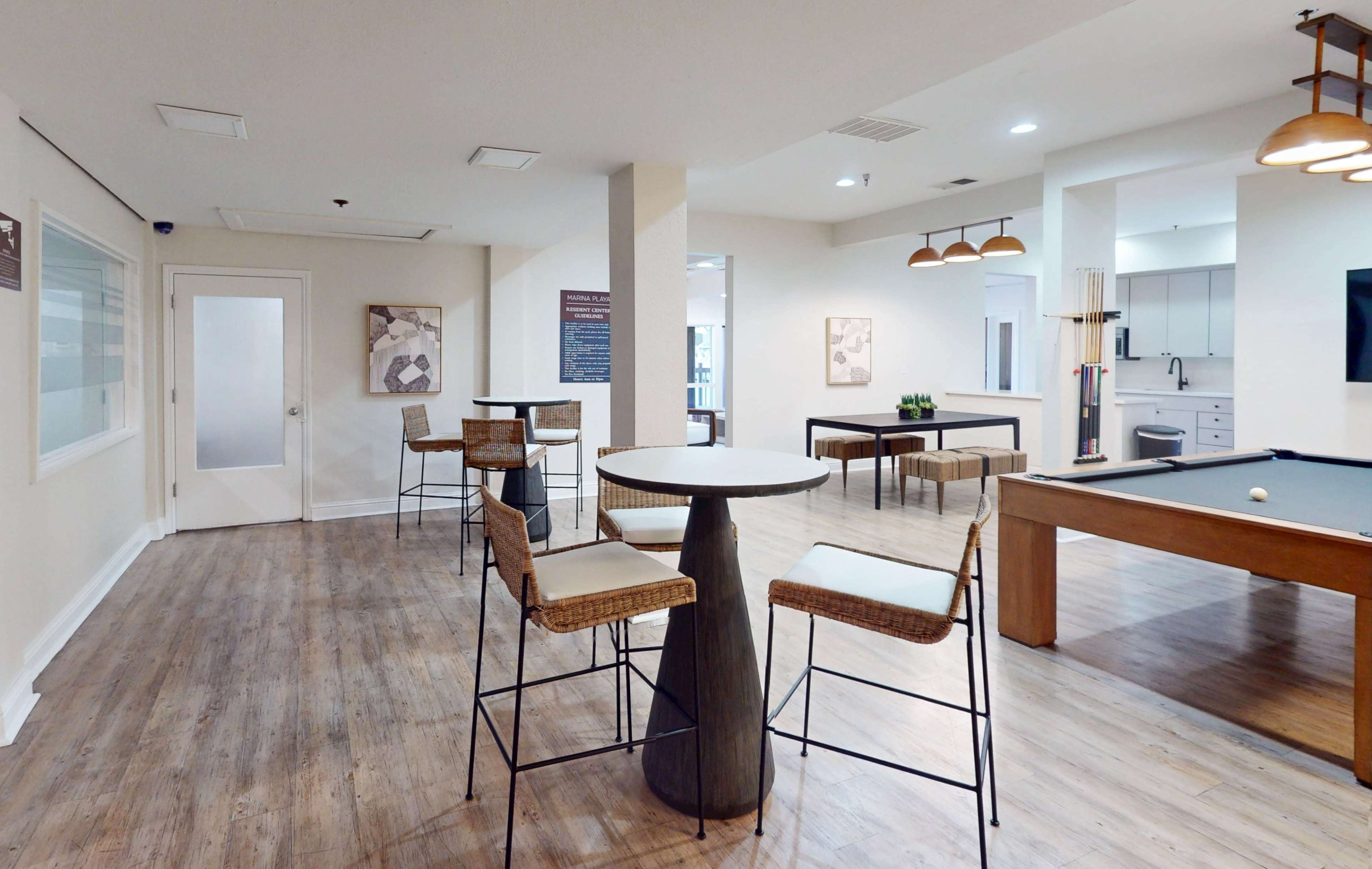 The image shows a modern communal space with a pool table, seating areas, and a kitchenette in a well-lit environment.