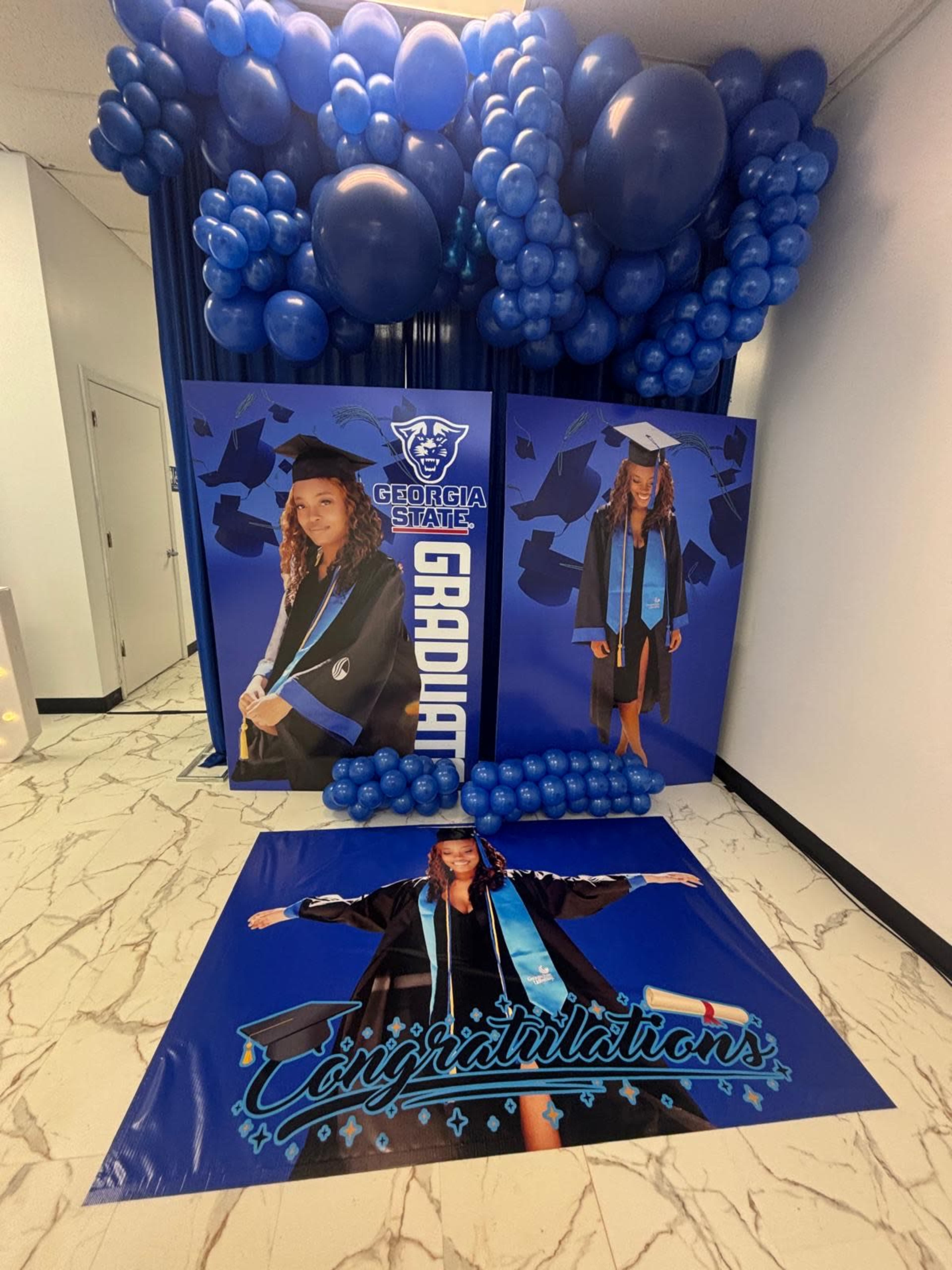 The image shows a decorated space for a graduation celebration, featuring blue balloons, posters of a graduate from Georgia State University, and a congratulatory message.
