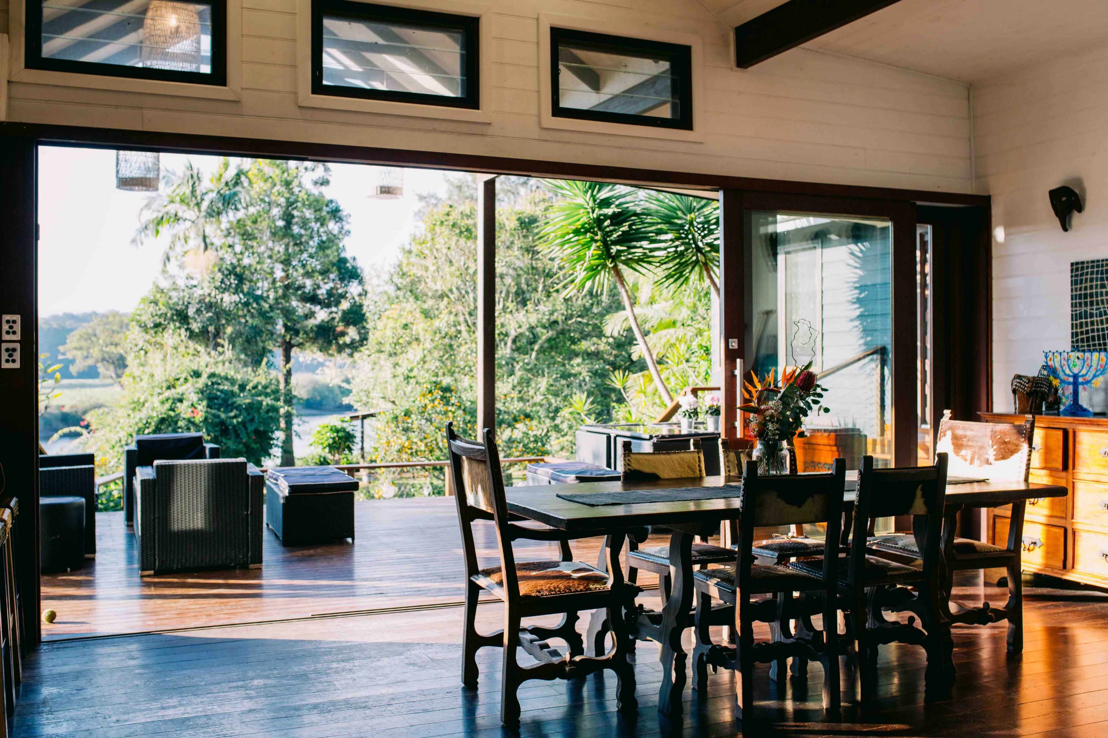 A dining area with a wooden table and chairs overlooks a patio surrounded by greenery through large glass doors.
