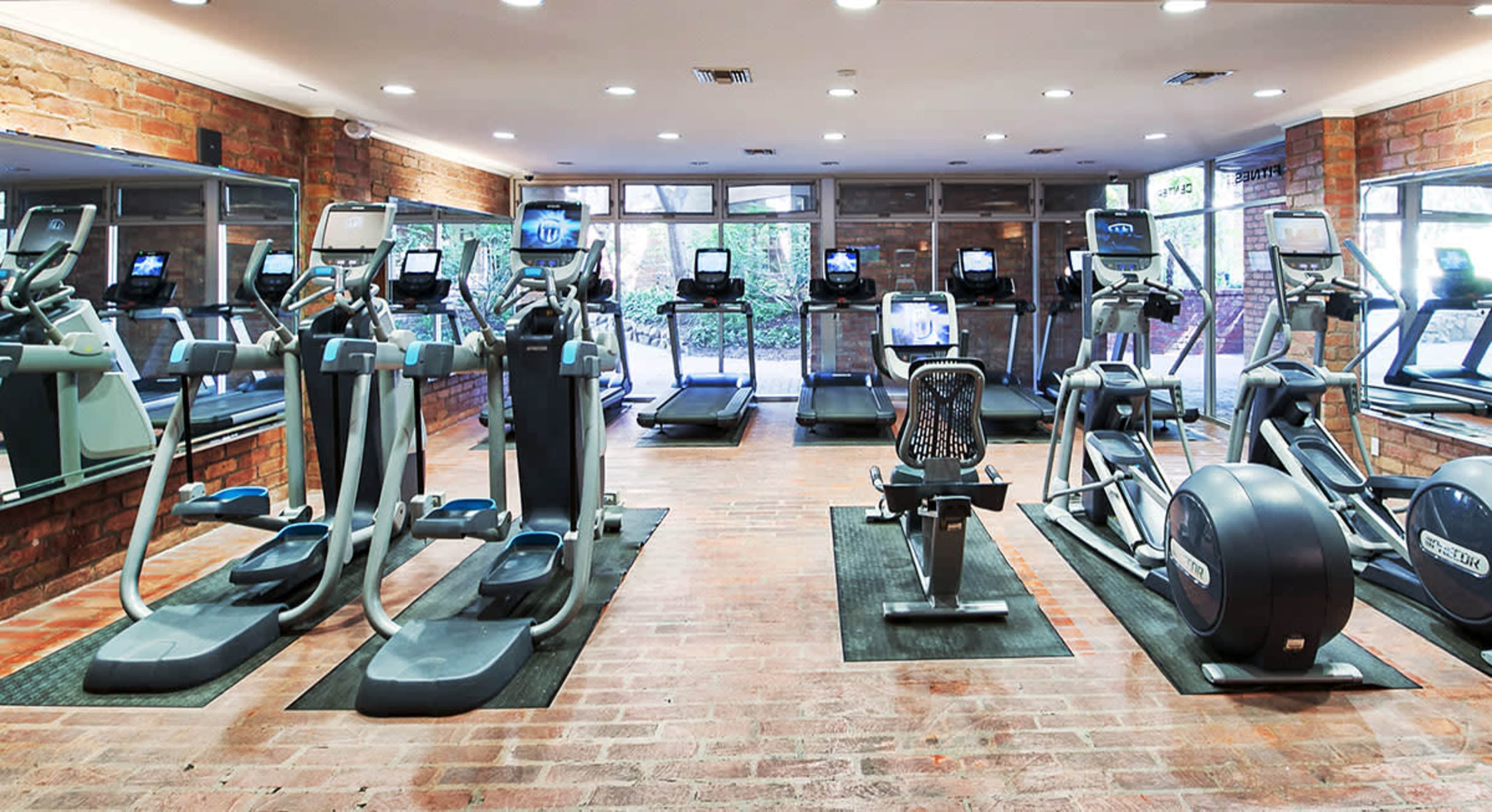 The image shows a gym interior equipped with various exercise machines, including treadmills, elliptical trainers, and stationary bikes, set against a backdrop of exposed brick walls and large windows.