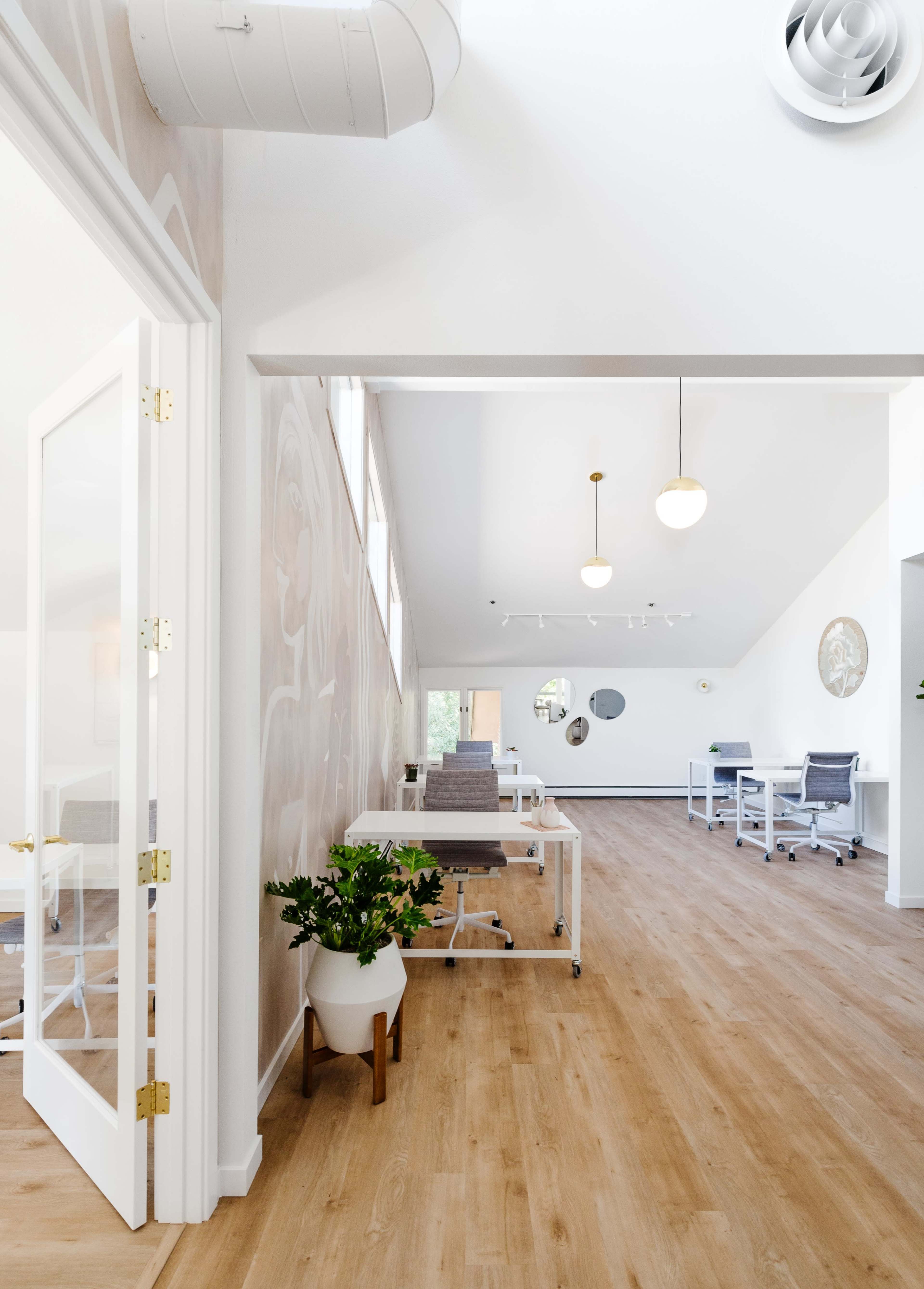 A modern, open office space with light wood flooring, minimalist furniture, and large windows allowing natural light.