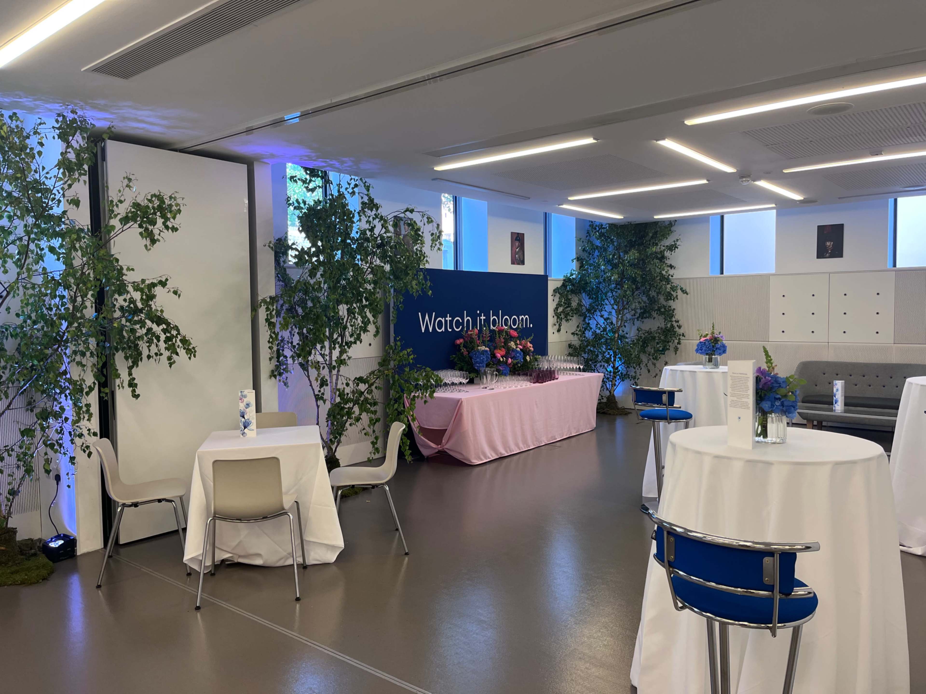 The image shows a modern event space with tables covered in white linens, a pink tablecloth display at the back, and decorative plants placed around the area.
